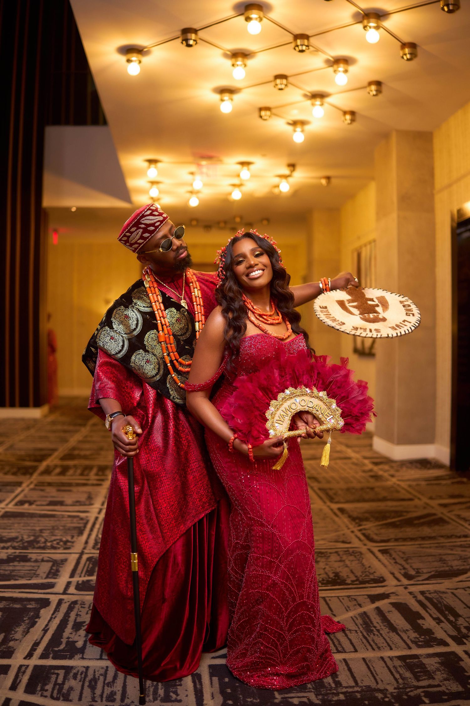 Two people in elegant red dresses dance and twirl together in a warmly lit hotel hallway with patterned carpeting.