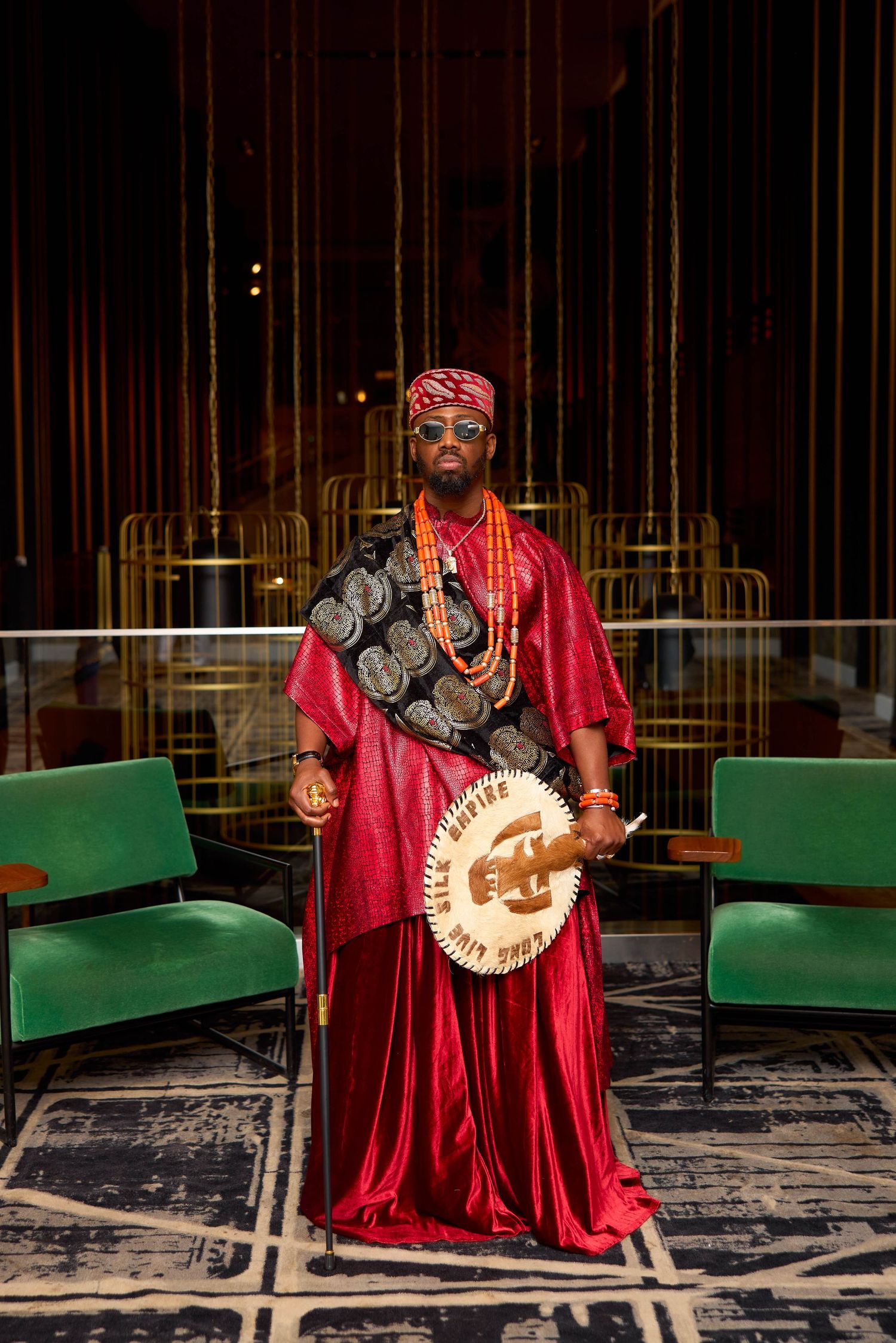 Figure in flowing red robes stands dramatically in a dark space with green seating and ornate decorative elements.