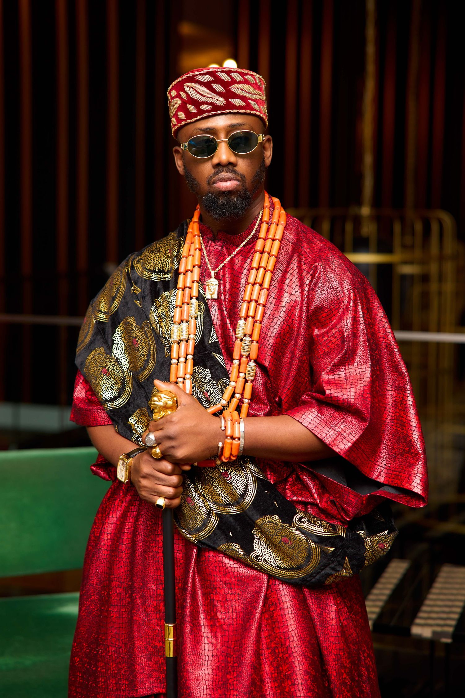 Person wearing traditional Nigerian attire in red agbada and gold beaded necklaces poses with stylish sunglasses.
