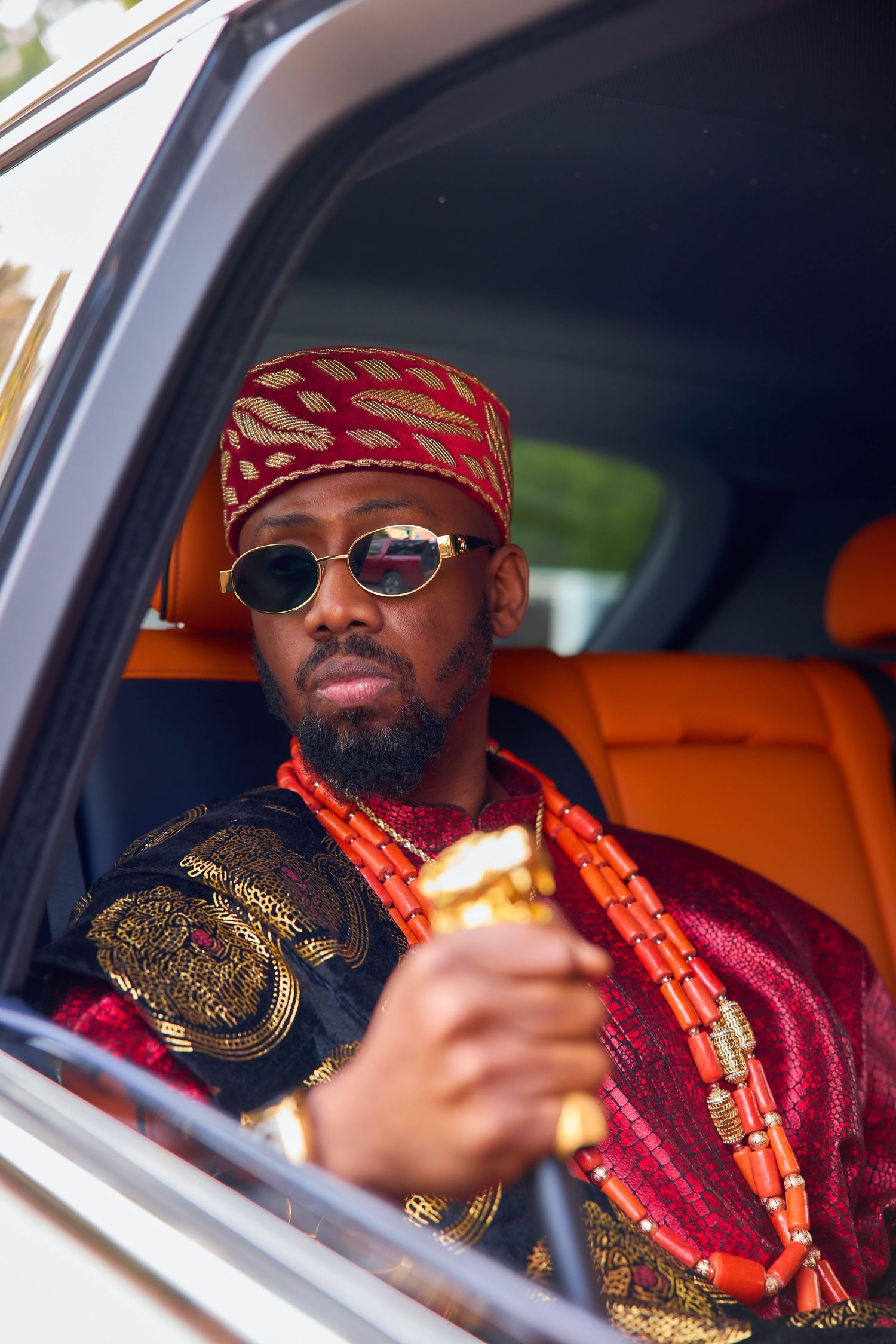 A person in traditional African attire with red beads and headwrap sits in a luxury car with orange leather seats.