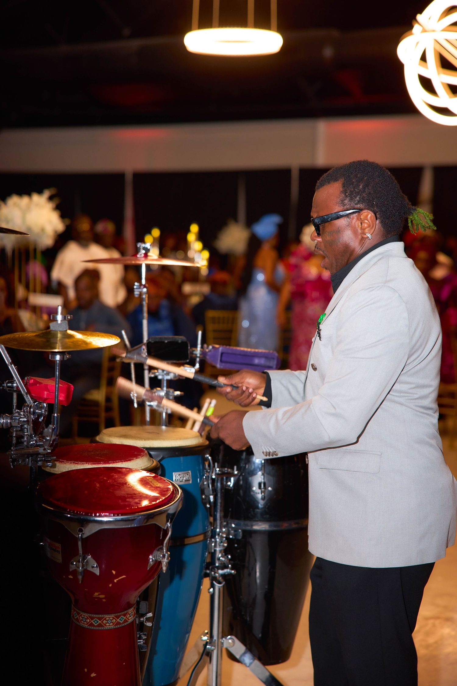 A musician in a white blazer plays percussion instruments at a lively indoor event with colorful lighting.
