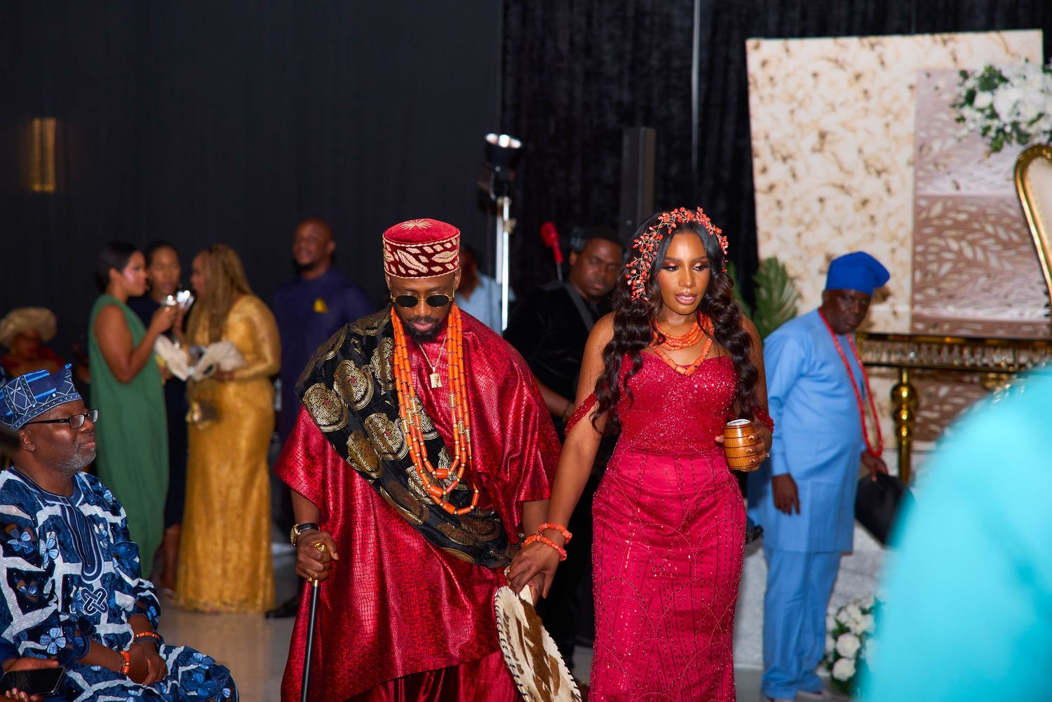 A group of people gather at an indoor ceremony wearing colorful traditional attire in red, blue and gold.