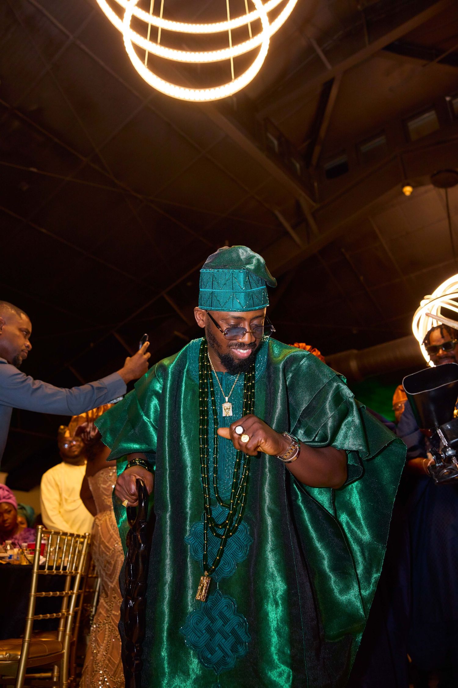 A person wearing emerald green traditional robes dances energetically at an indoor celebration.