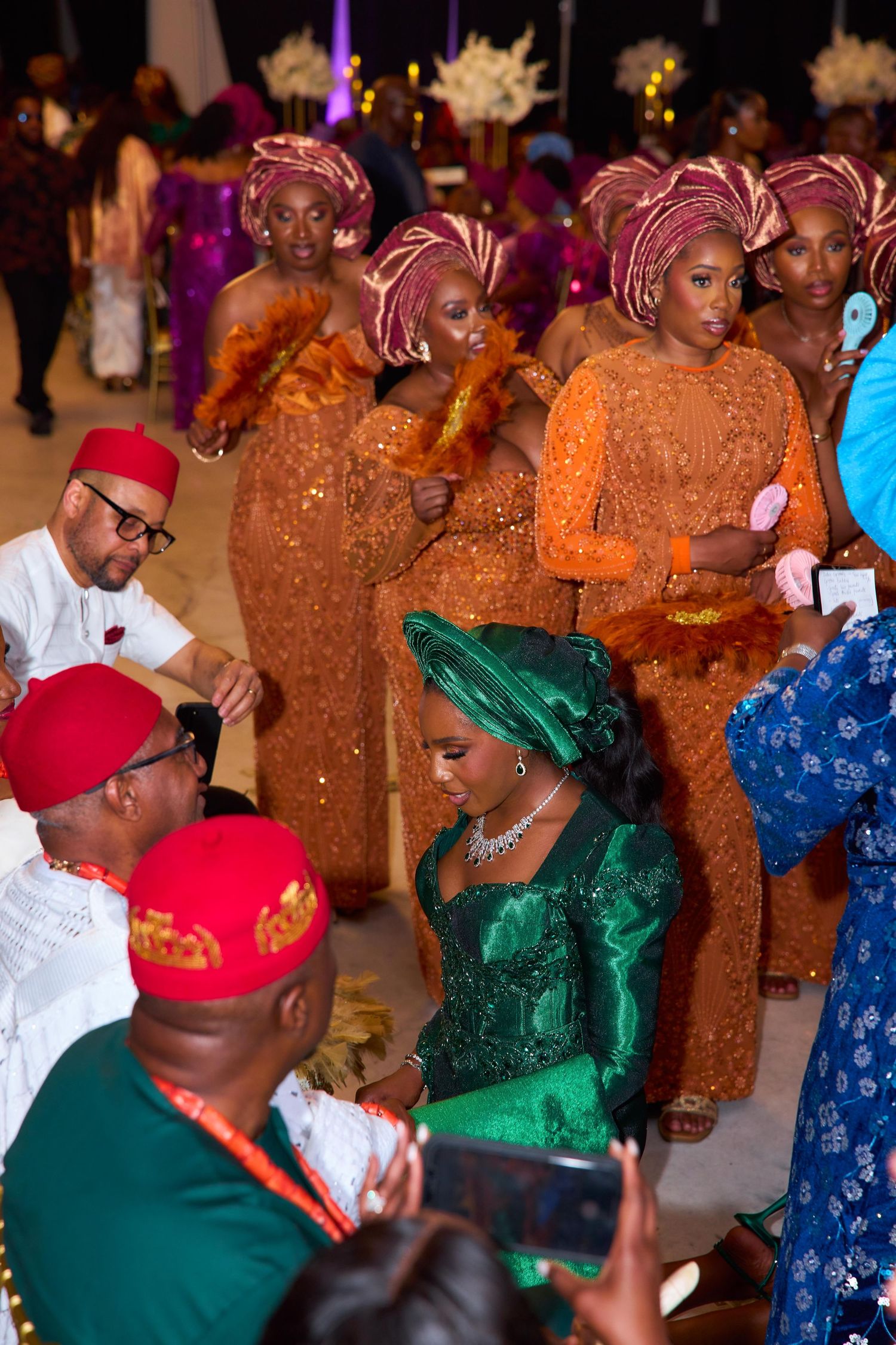 Celebrants in vibrant traditional Nigerian attire dancing at a formal event with emerald and orange garments.