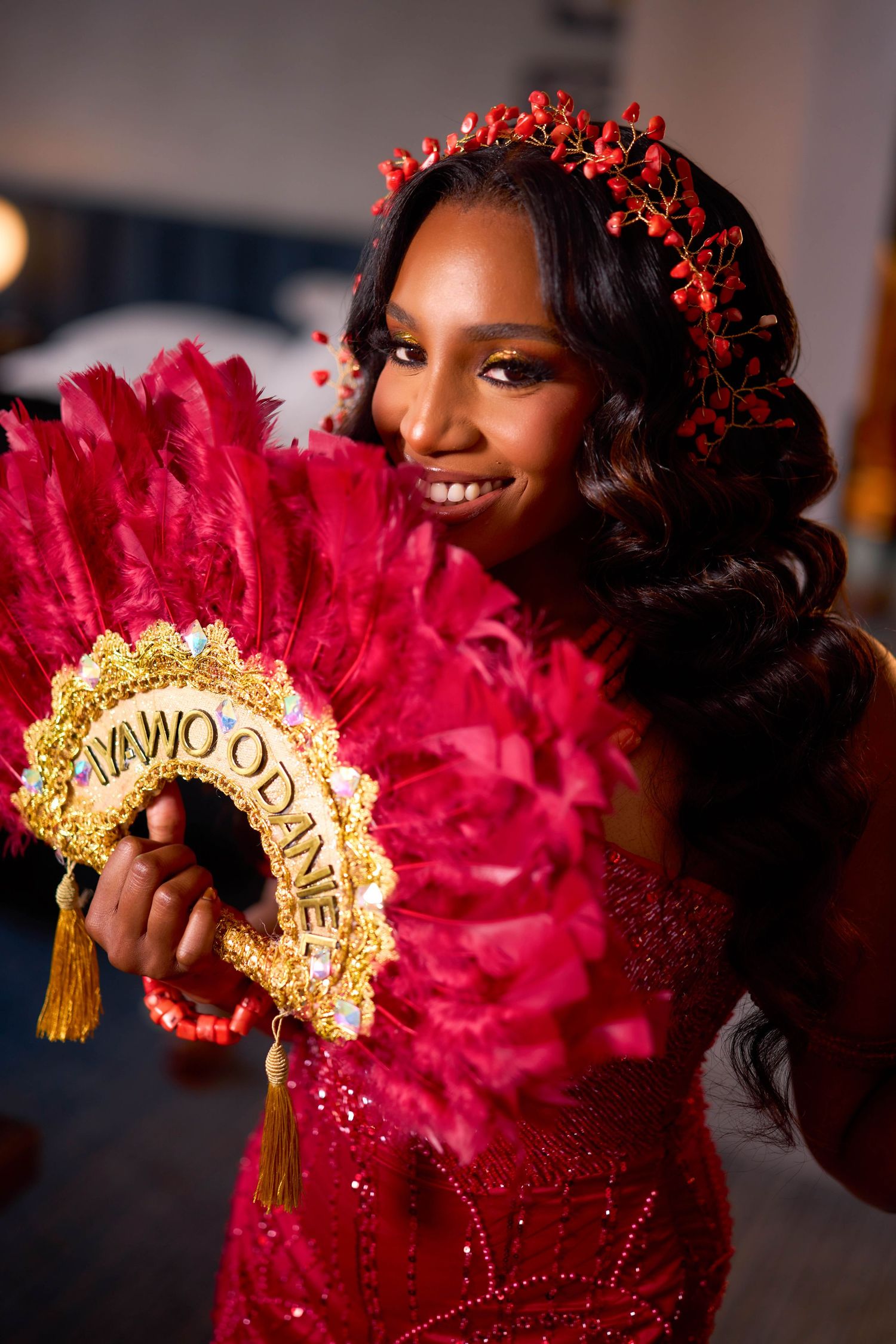 A woman in a red gown with a matching floral crown holds a decorative gold and red fan against a dark background.