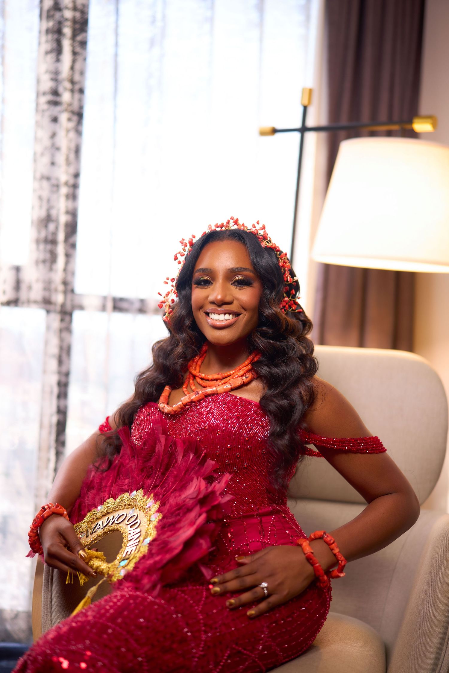 Person wearing a sparkling red dress poses elegantly on a cream colored chair in a well-lit room.
