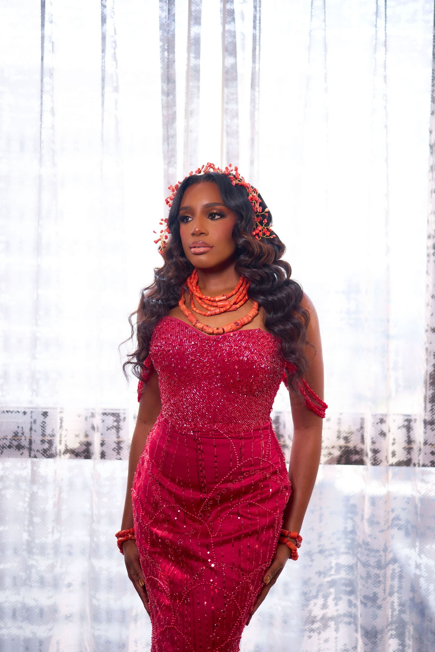 Person in a sparkling red evening gown poses elegantly against a light curtained backdrop.