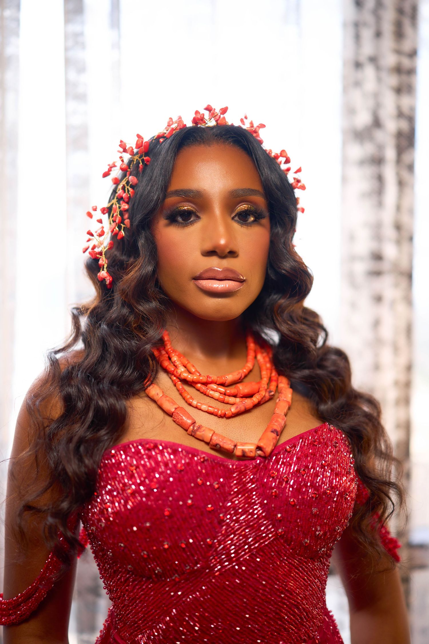 A person in a sparkly red gown with long dark curls and red floral accessories poses in front of a light background.