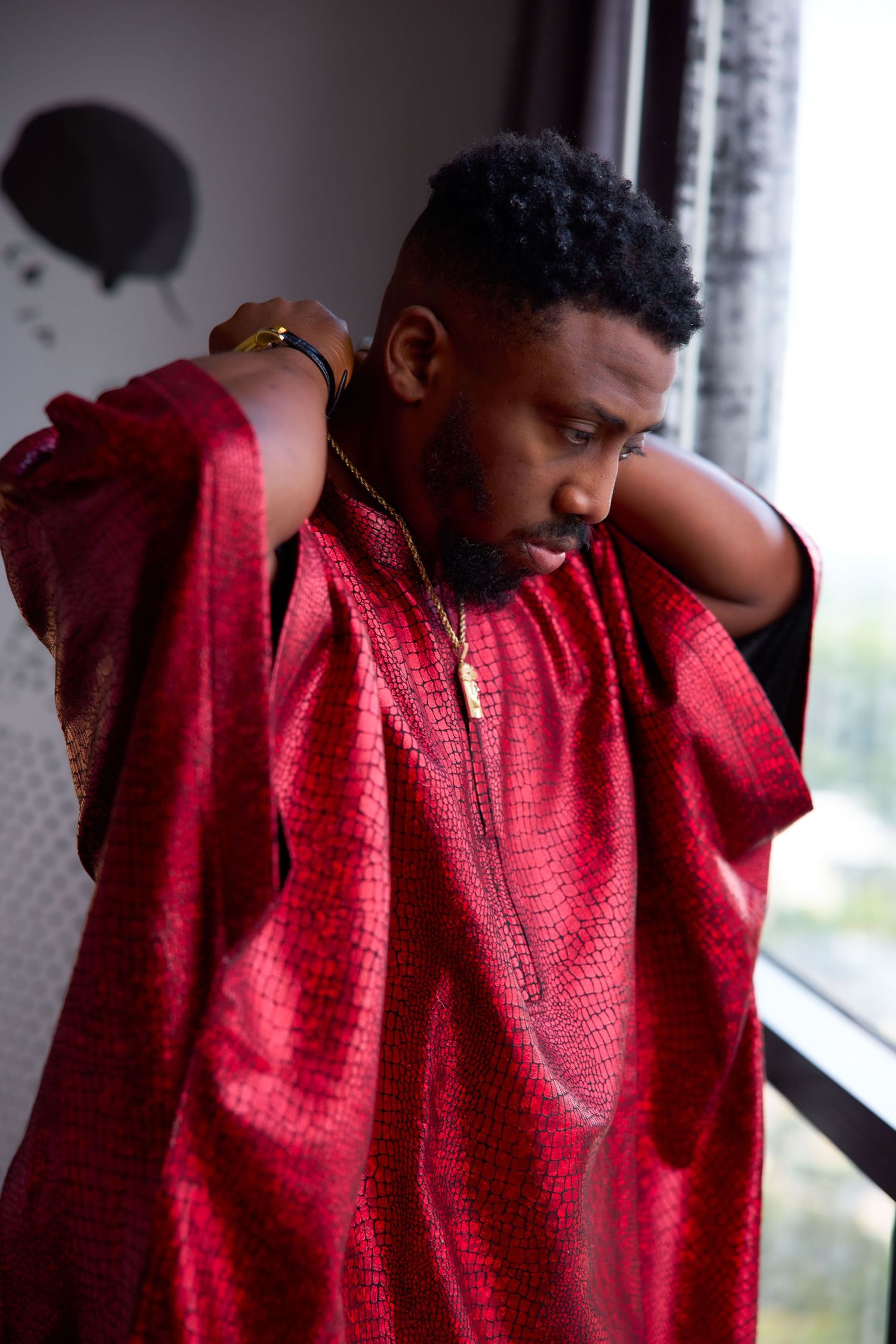 A person in traditional red attire stands by a window in a contemplative pose with natural lighting.