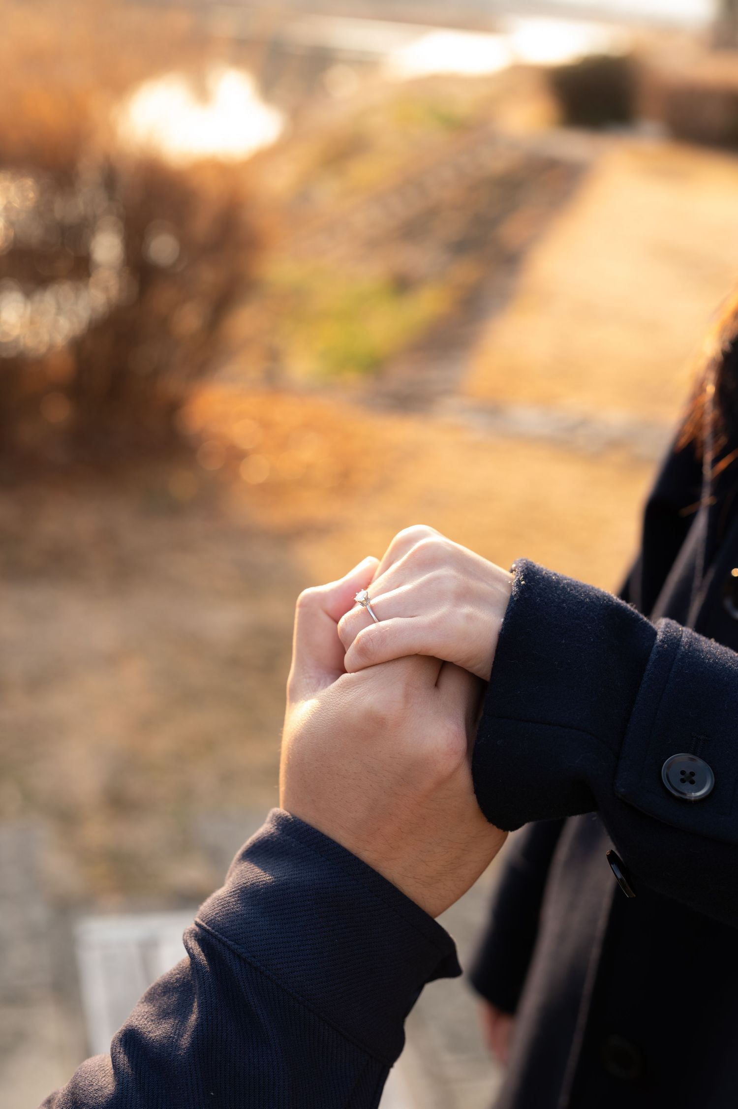 Close-up of couple holding hands with engagement ring visible at golden sunset outdoors.