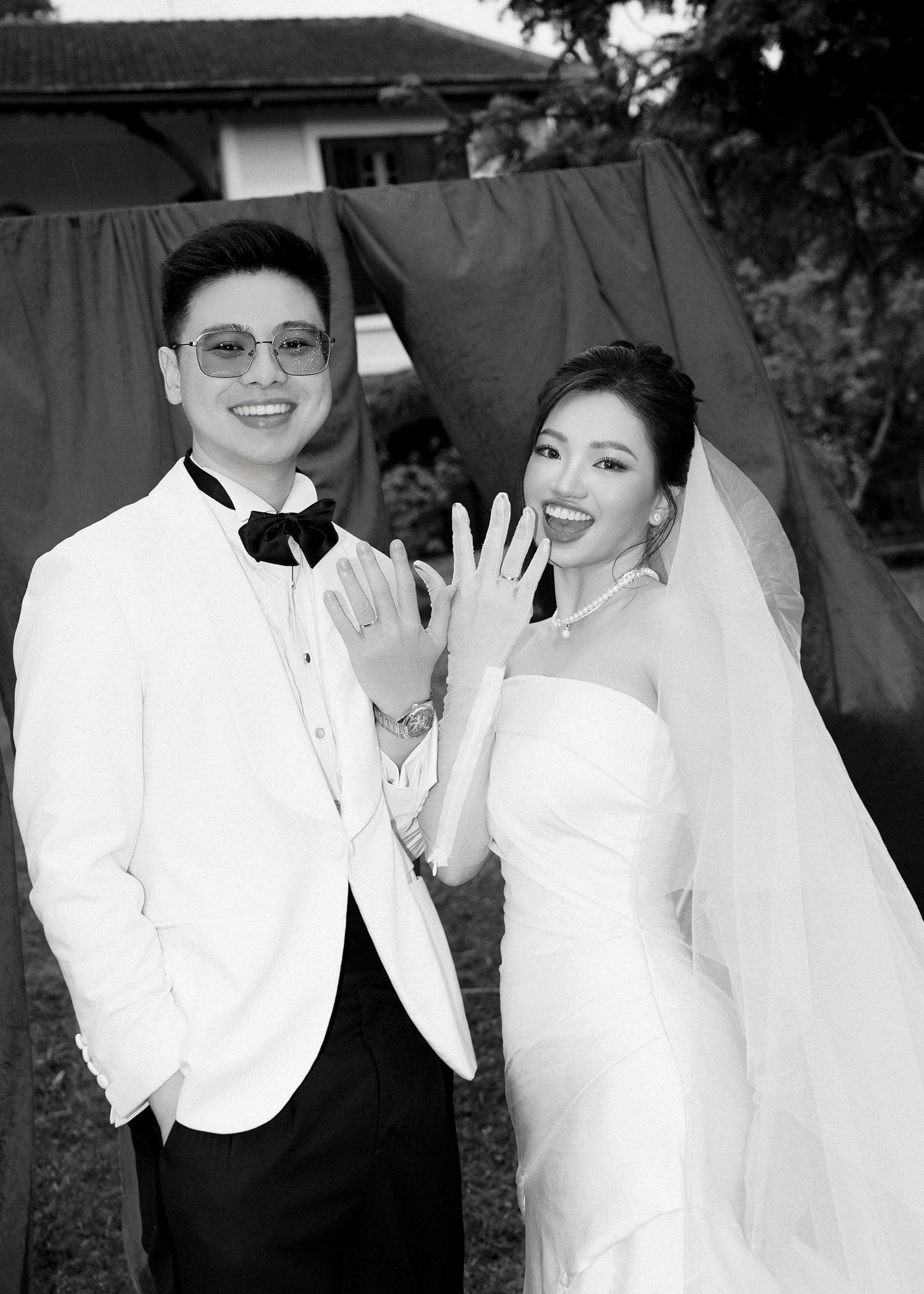 Smiling Asian newlyweds show off wedding rings in black and white photo with draped backdrop behind them.