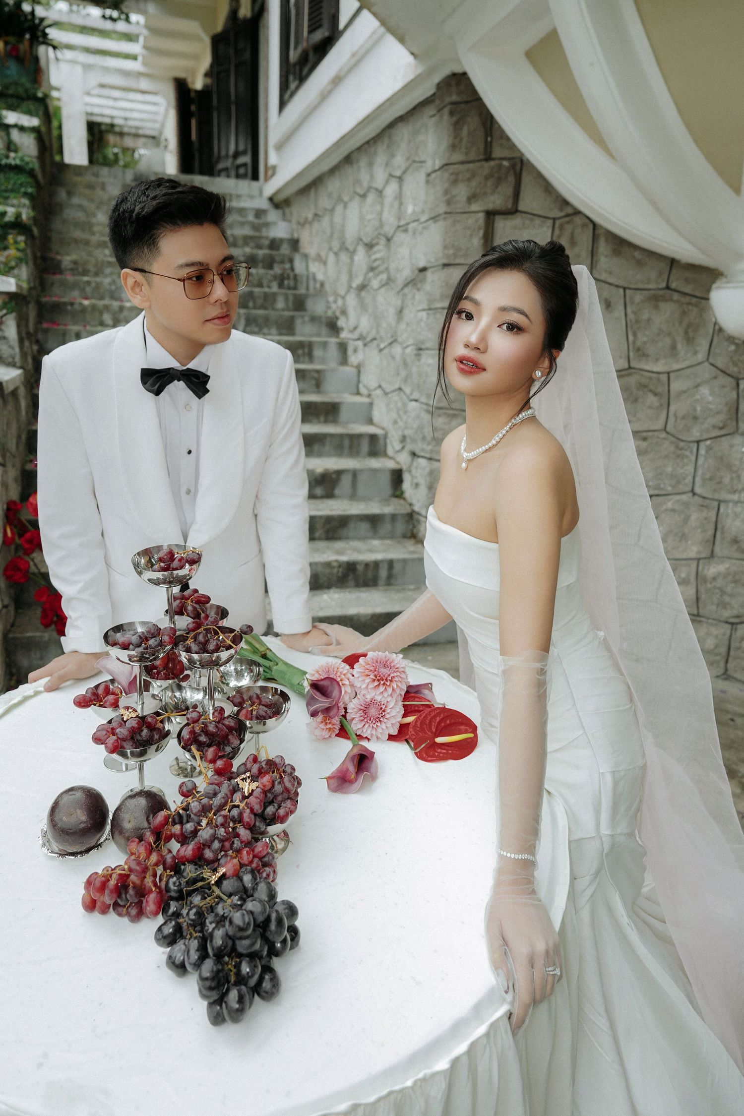 Elegant Asian couple in wedding attire pose at a table adorned with red grapes and flowers near stone steps.
