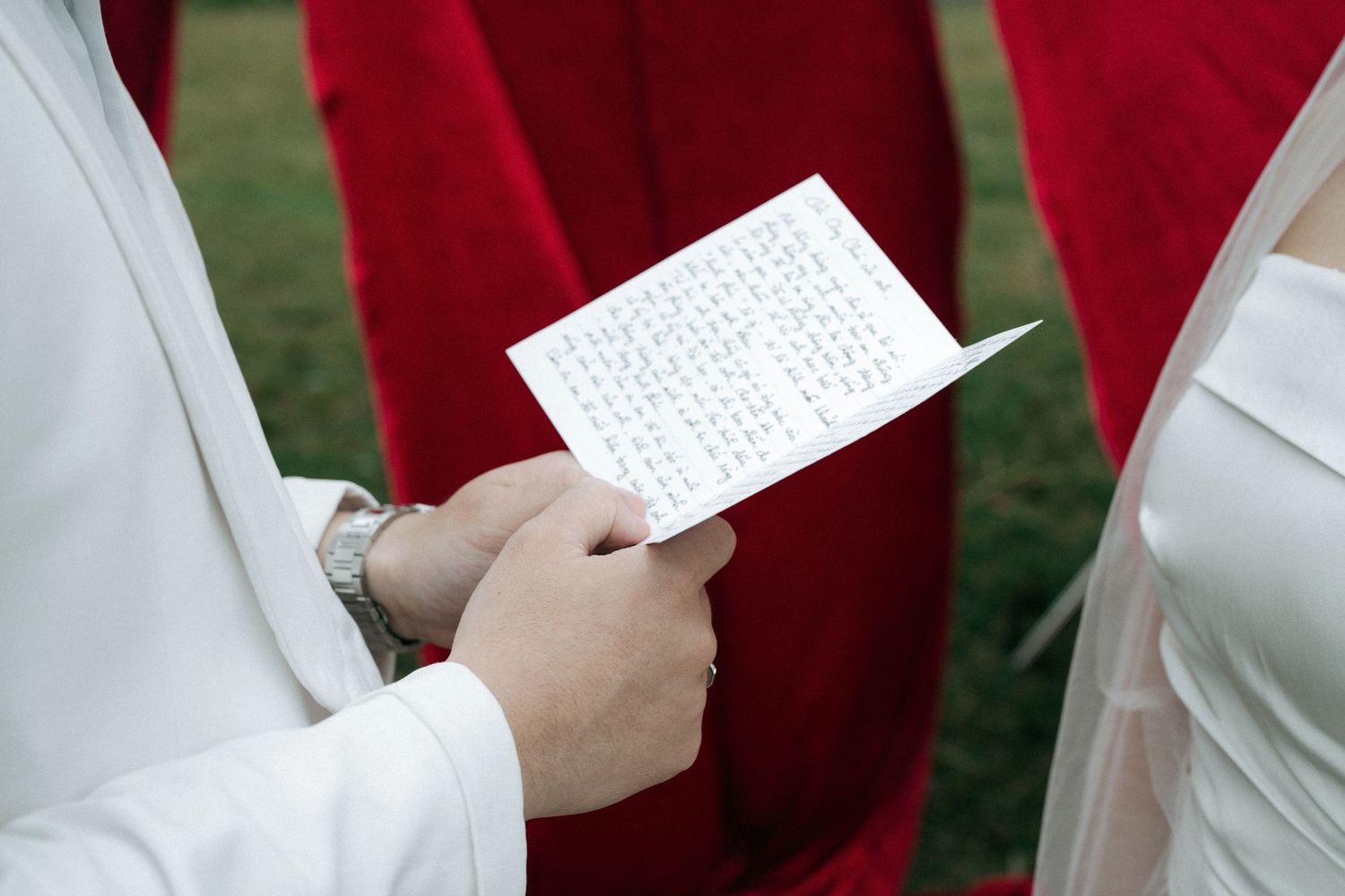 Close-up of groom holding handwritten wedding vows card with bride in white gown nearby.