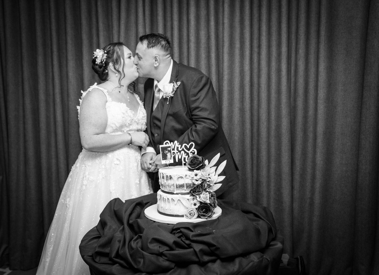 Black and white photo of a couple sharing a kiss behind their multi-tiered wedding cake at their reception.