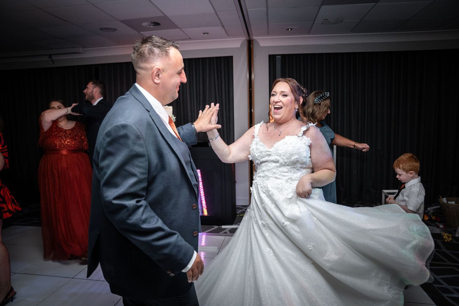 Wedding guests dancing and celebrating at an elegant indoor reception venue with joyful energy on the dance floor.