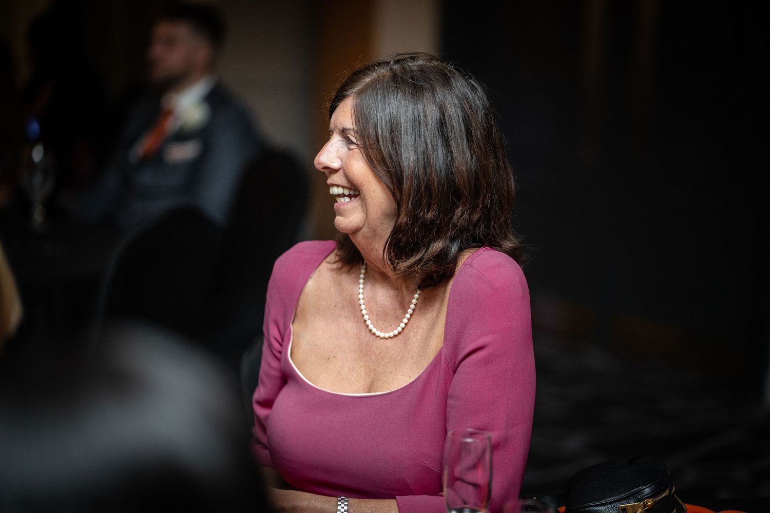 Woman in burgundy top with pearl necklace smiling during indoor event.