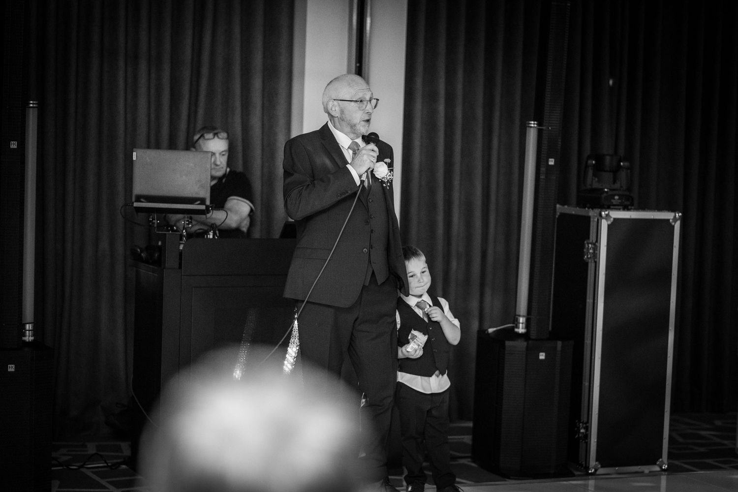 A black and white photograph shows a performer singing onstage with curtains and stage lighting visible behind them.