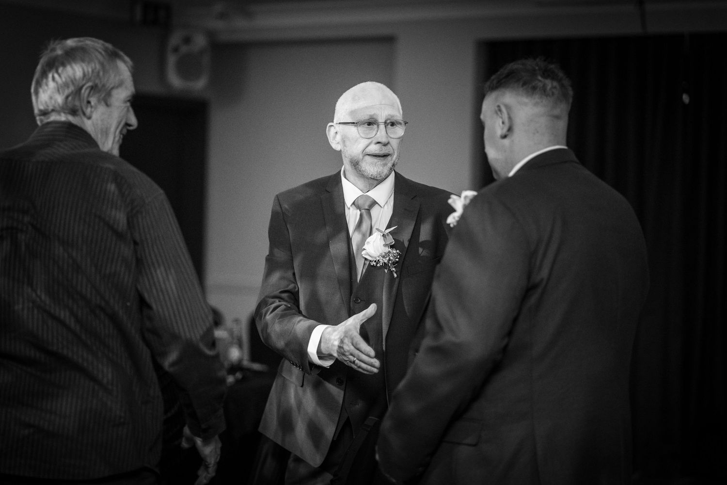A black and white photograph shows three men in suits having an animated conversation at an event.