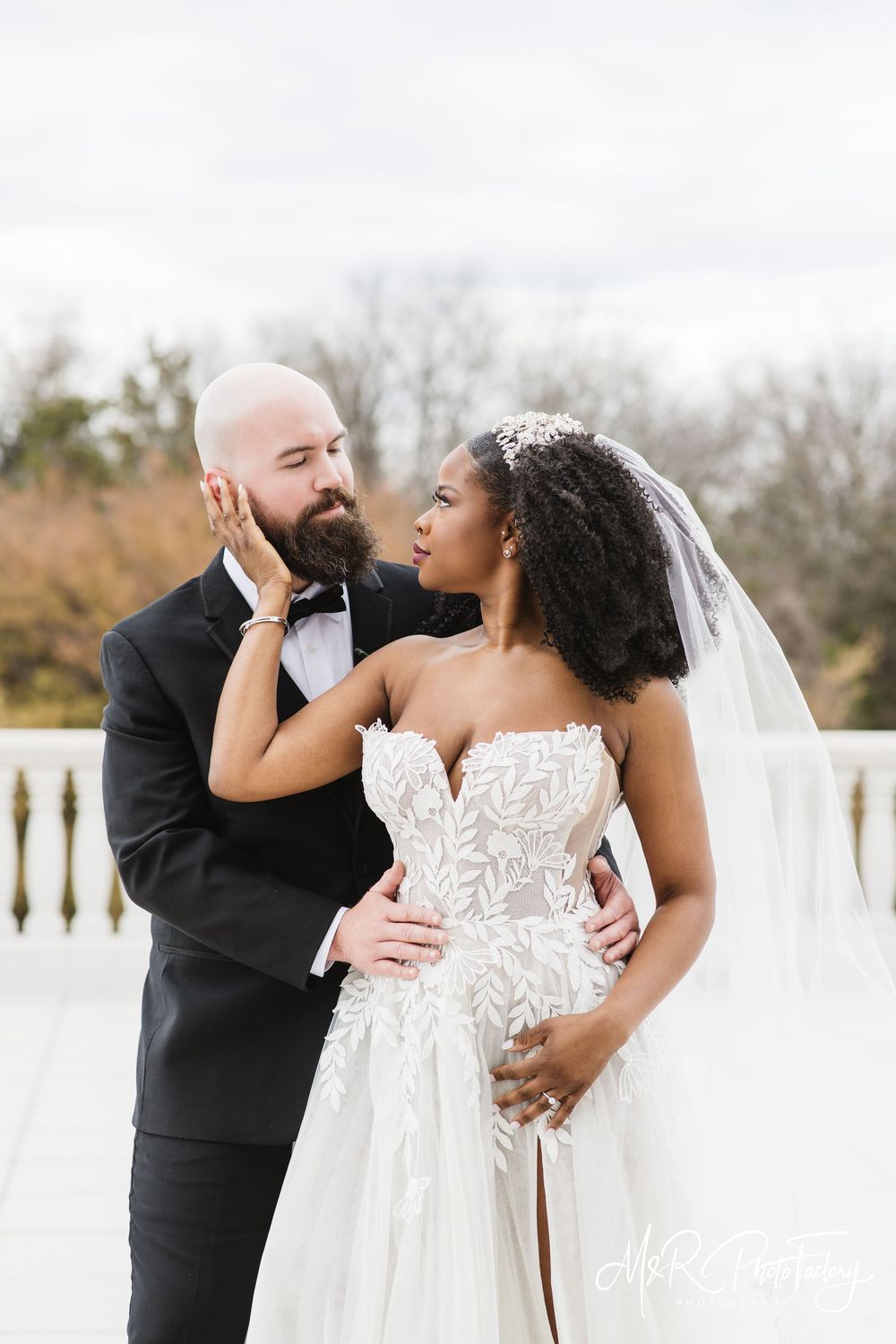 Interracial wedding couple sharing tender moment outdoors, bride in floral lace ballgown with veil.
