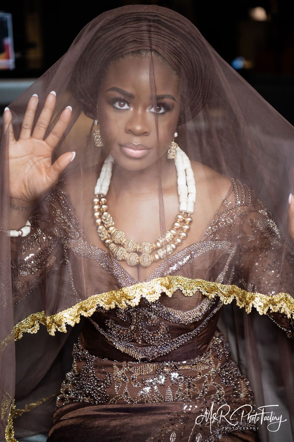 Stunning Black bride in brown traditional African wedding attire waving through sheer golden veil.