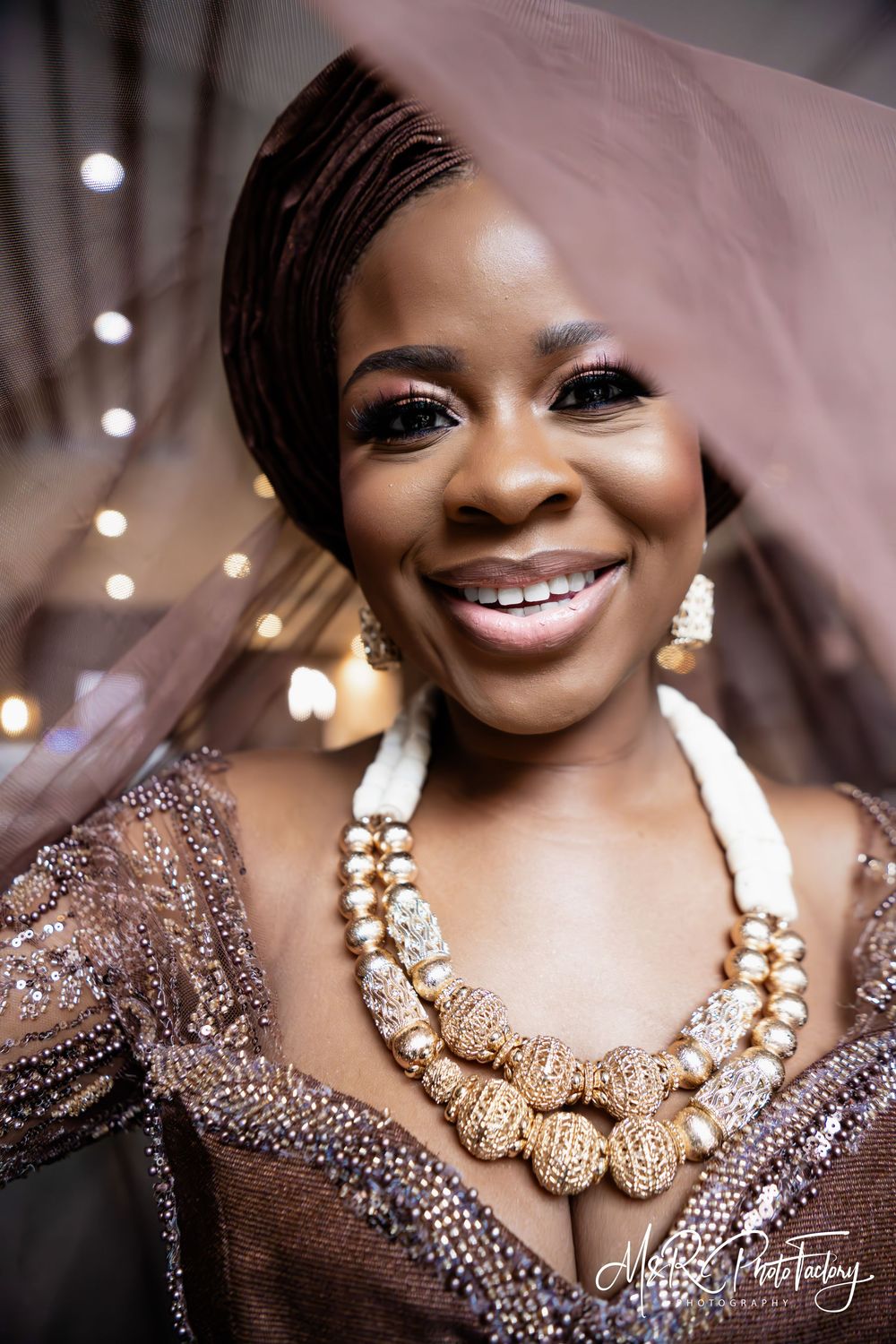 Glowing Black bride in brown African traditional wedding dress with gold beaded necklace smiling.