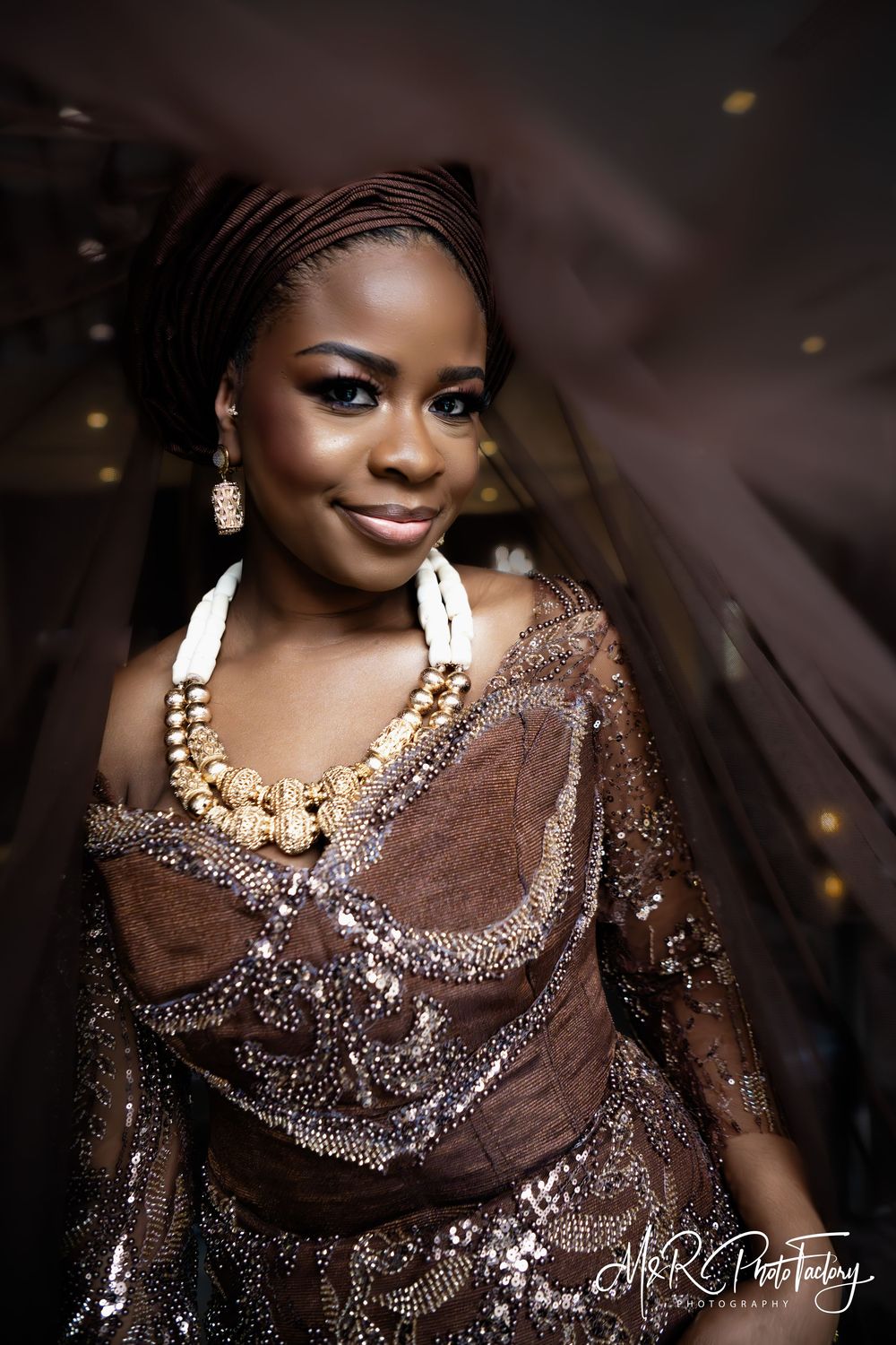 Beautiful African bride wearing brown traditional attire with gold jewelry and blue eye makeup.