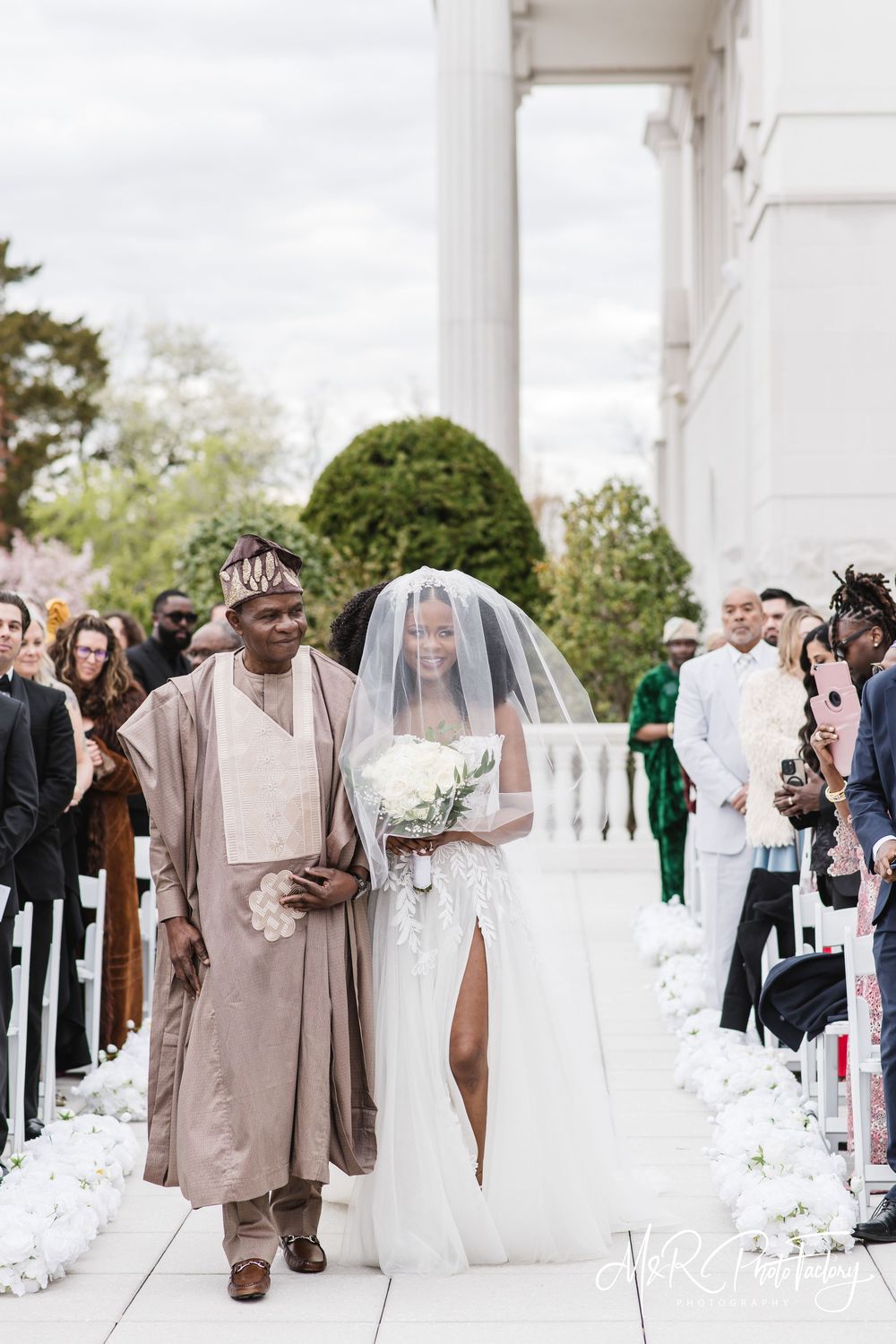Nigerian father in traditional attire walking bride down outdoor aisle lined with white flowers.