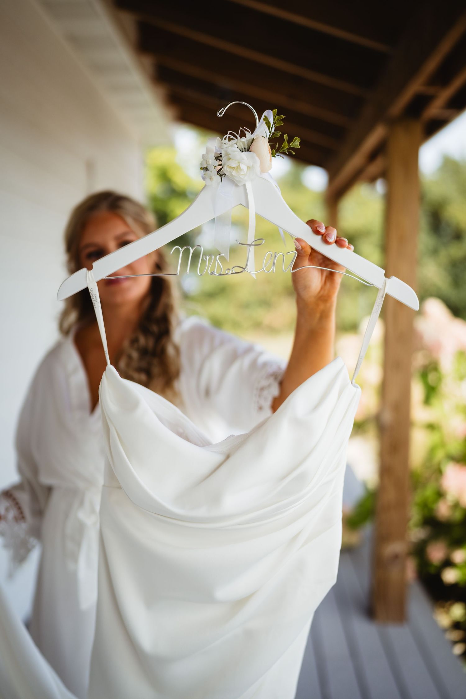 A white wedding dress hangs on a custom hanger under a wooden porch overhang in soft natural light.