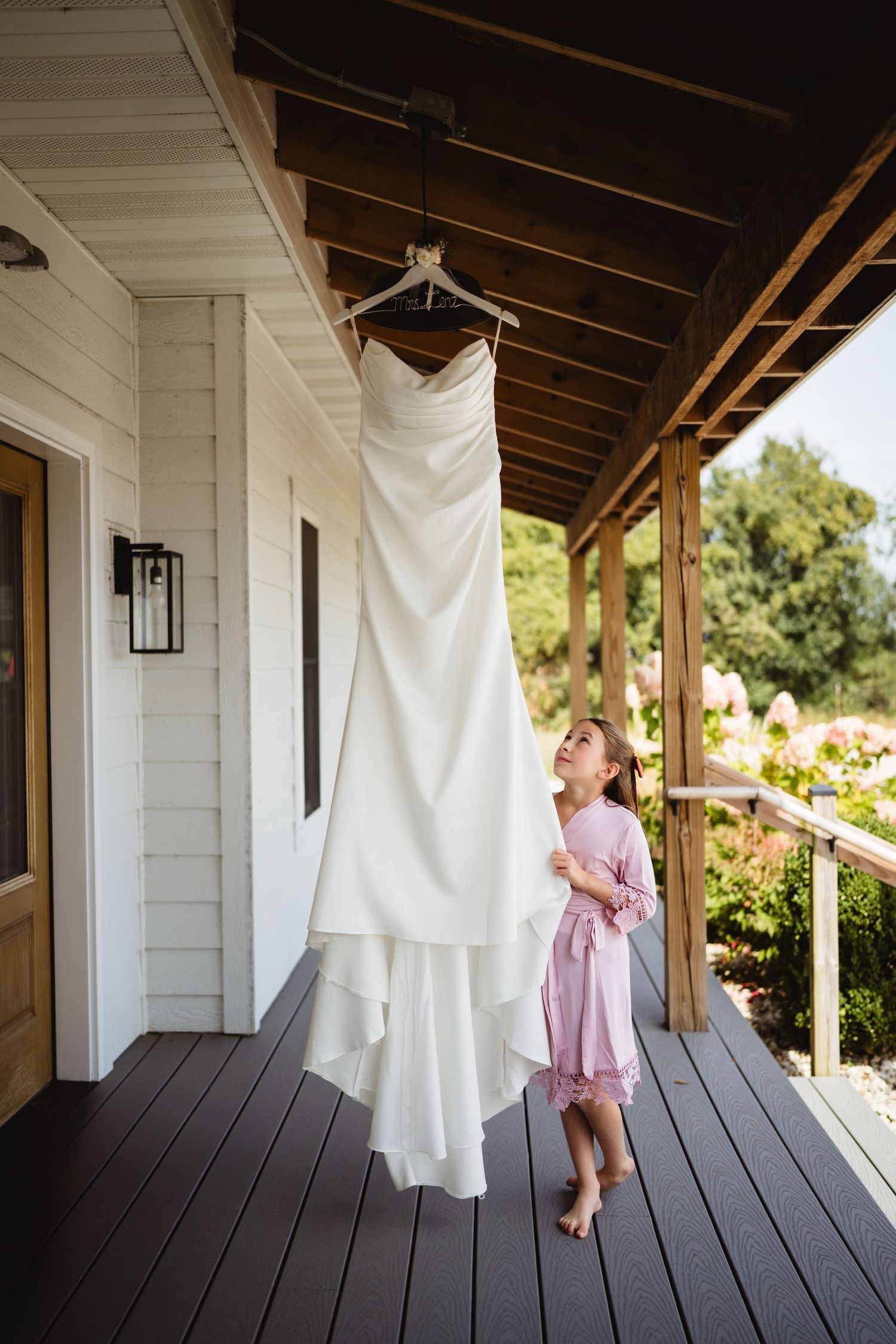 A white wedding dress hangs on a porch while a person in a pink dress looks up at it admiringly.