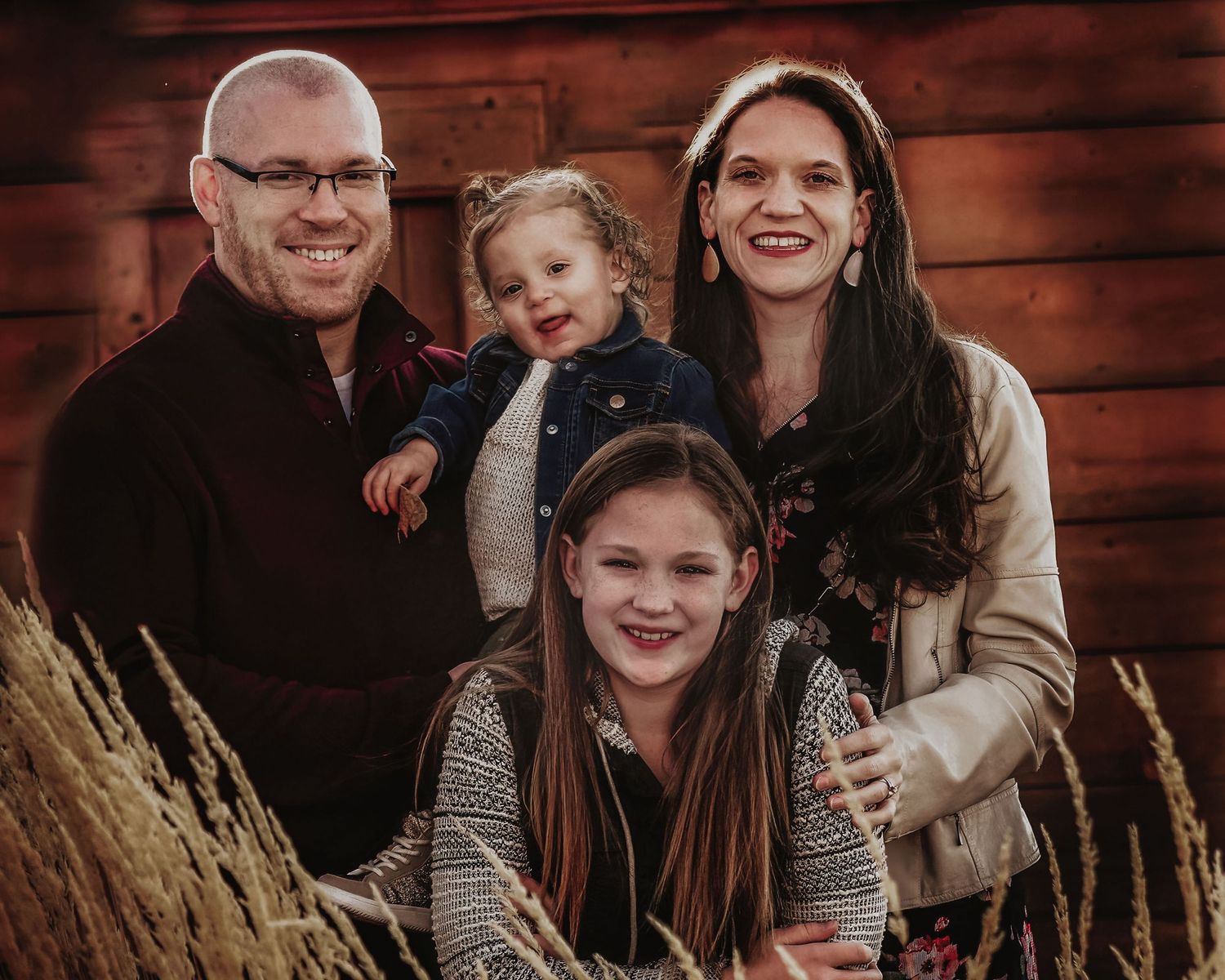 A warm family portrait of four people smiling together in front of a rustic wooden wall with dried wheat stalks.