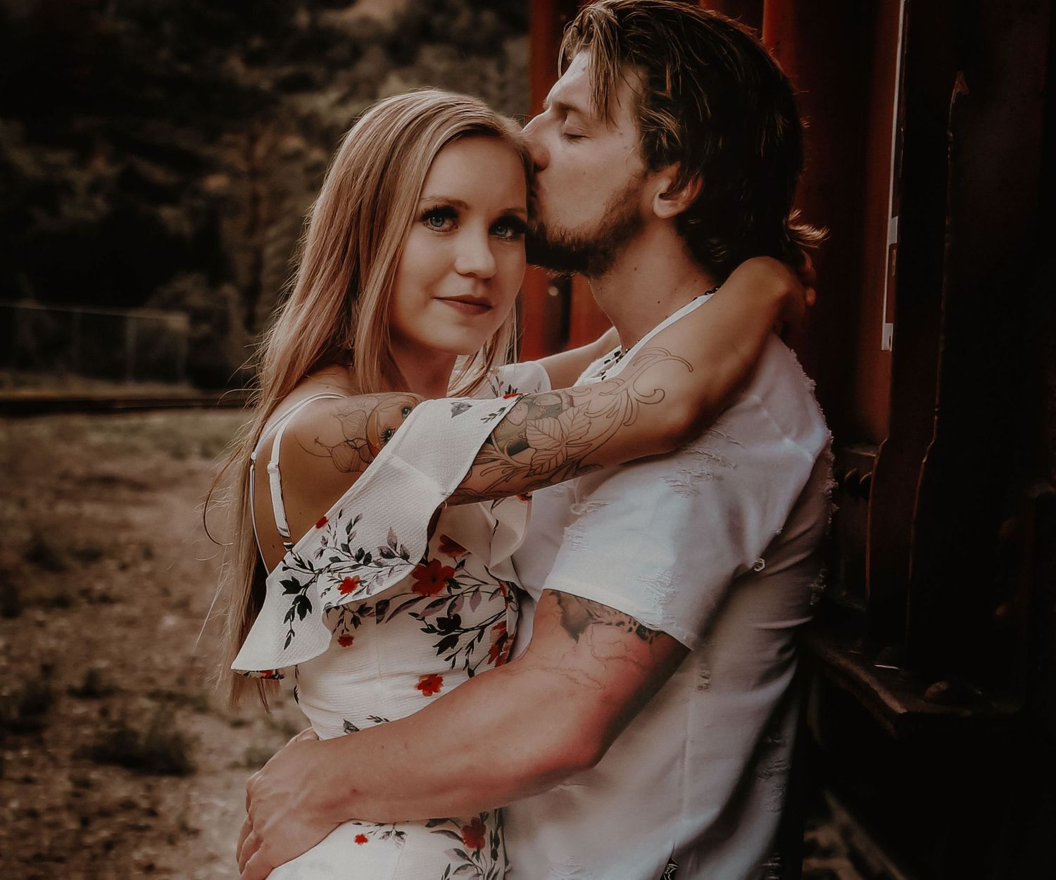 Romantic couple embraces outdoors in warm sunset light, with her wearing a white floral dress and him in casual attire.