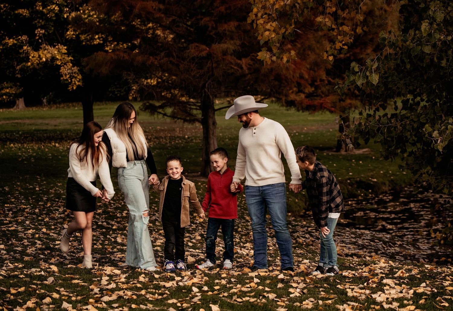 A group of people walking through a park covered in fallen autumn leaves during a fall photoshoot session.