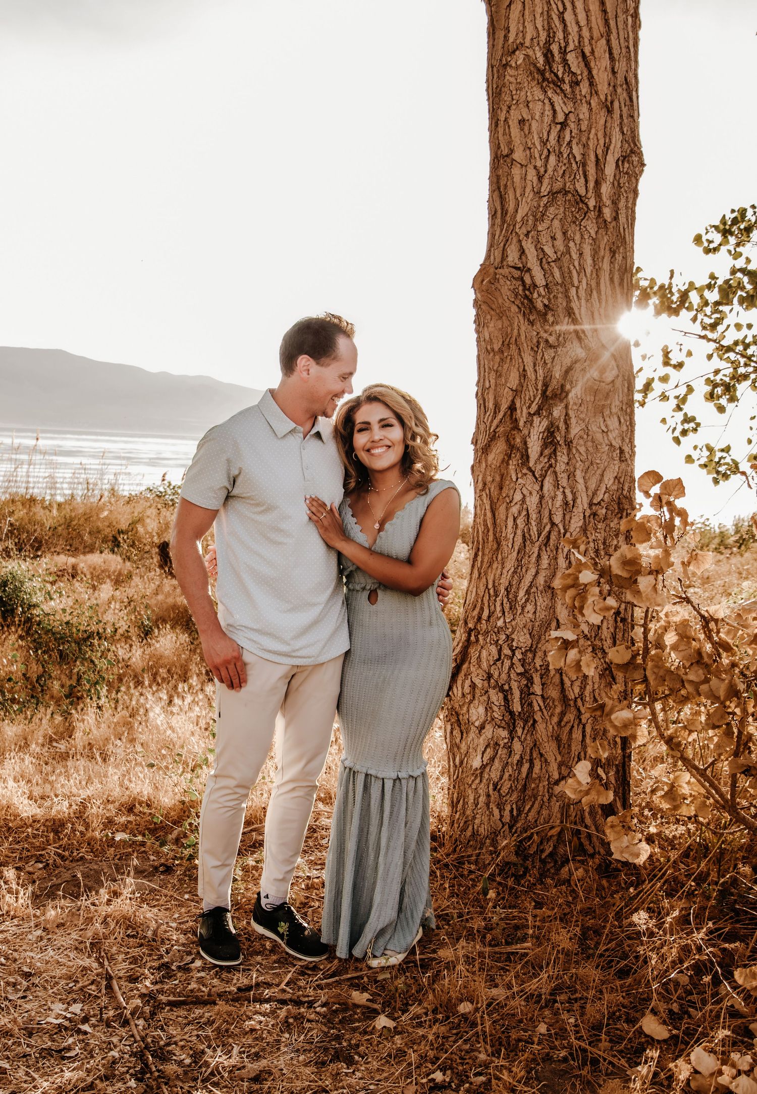 Romantic outdoor photo session at sunset with couple posing near large tree trunks in warm golden light.