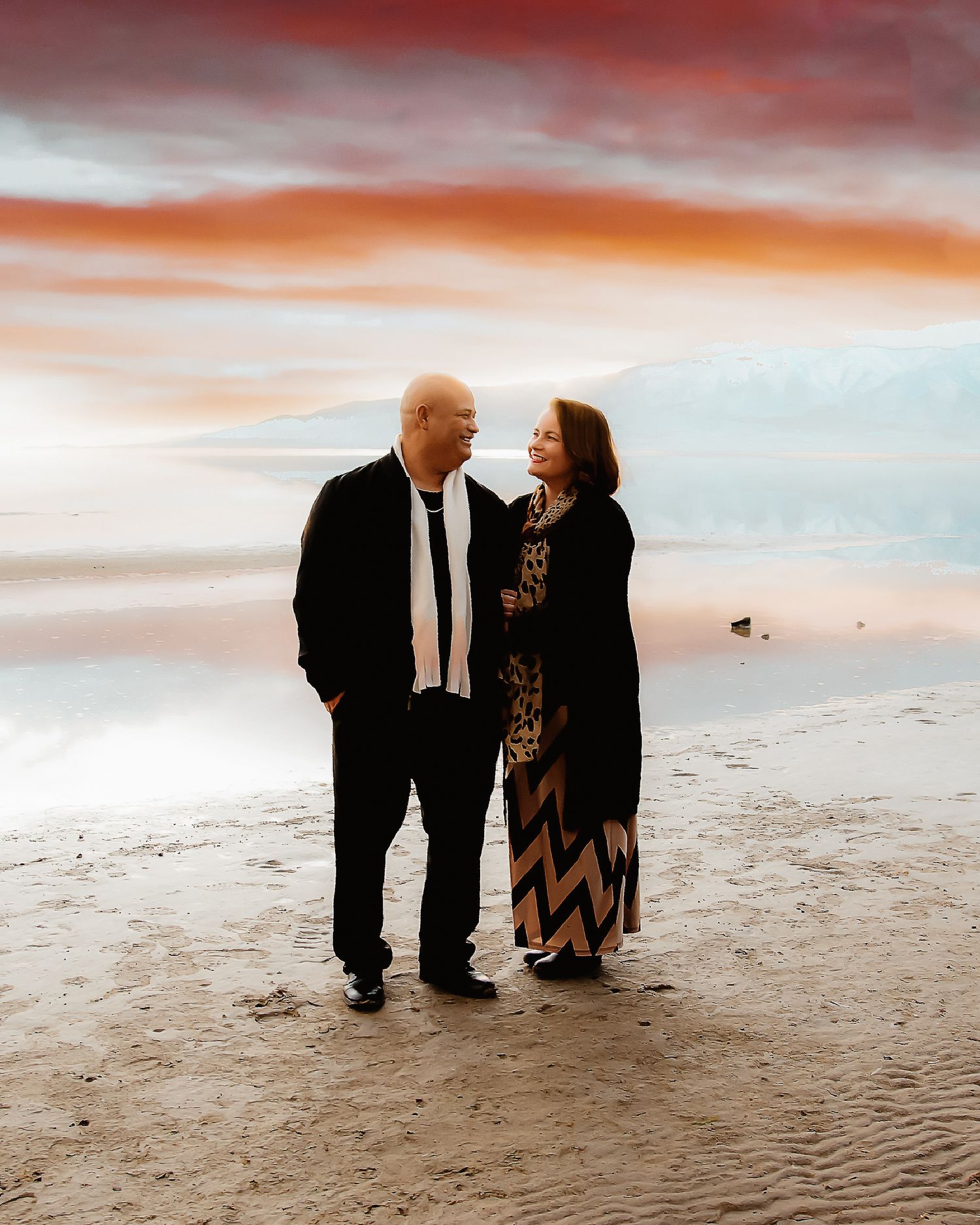 A couple stands together on a beach at sunset with dramatic orange and pink clouds in the sky.