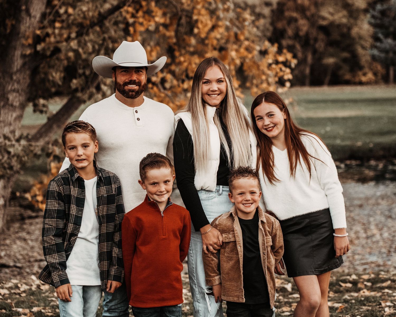 Family wearing coordinated fall outfits poses together outdoors among autumn leaves during sunset photo session.