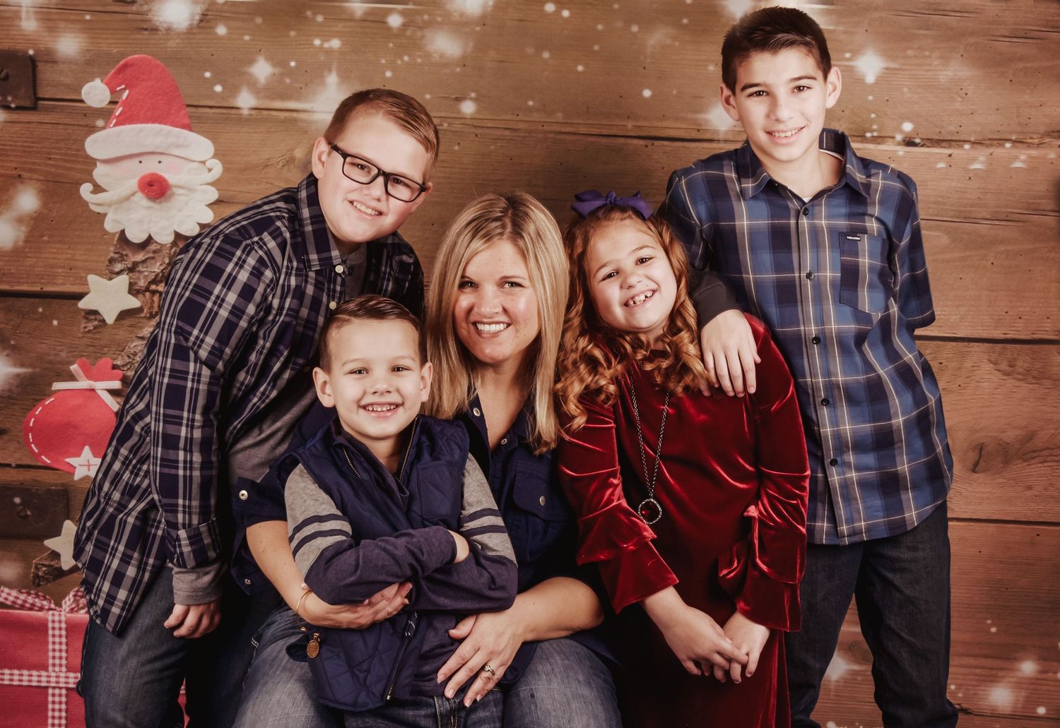 Family wearing coordinated red and navy outfits poses together against rustic wooden wall with falling snow effect.