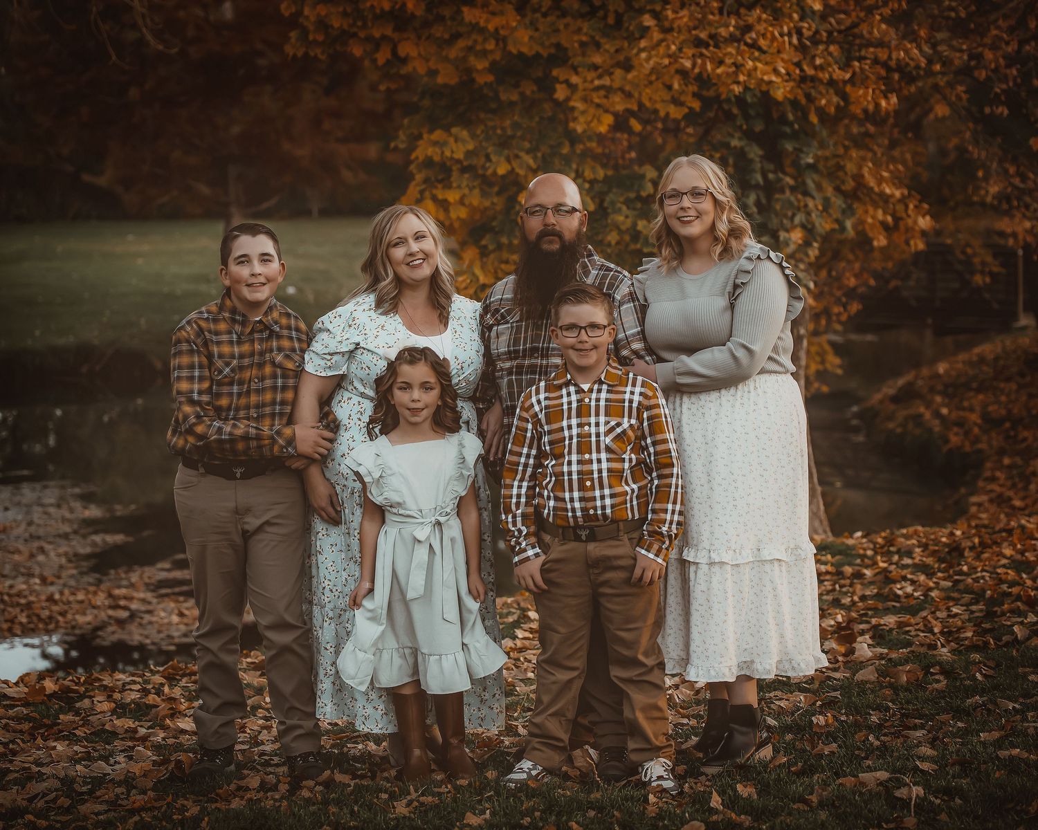 Family wearing coordinated fall outfits poses together on autumn ground covered in orange leaves during sunset.