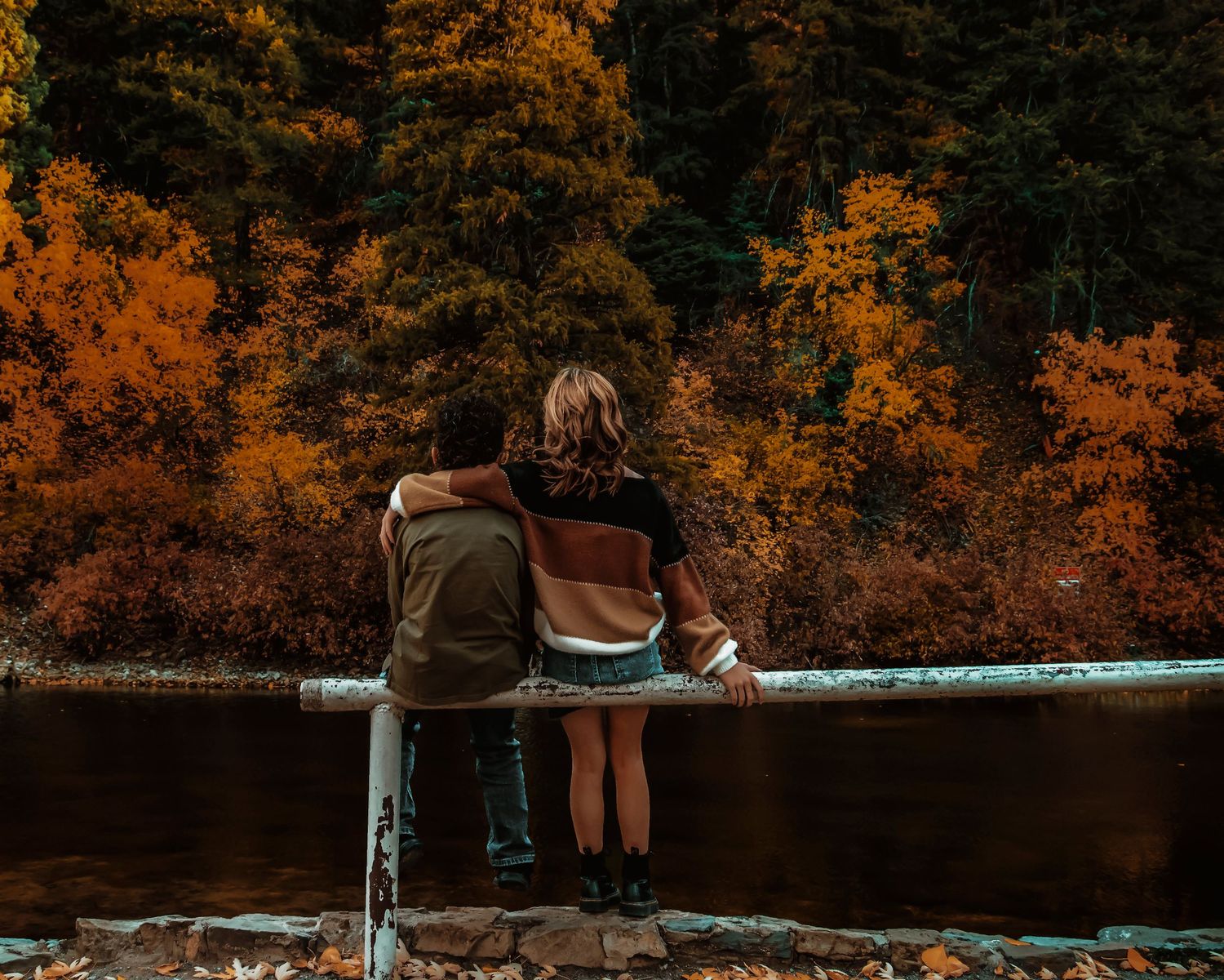 Two people sit together on stone wall overlooking lake surrounded by vibrant autumn foliage in forest.