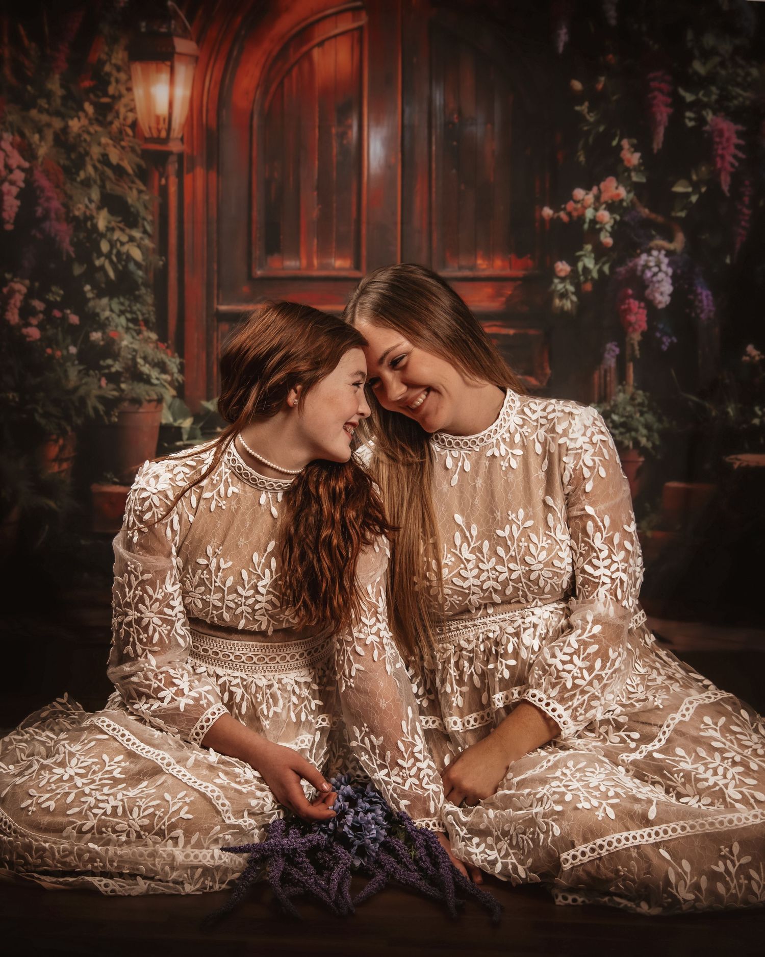 Two people in matching white lace dresses pose intimately in a romantic garden setting with moody lighting.
