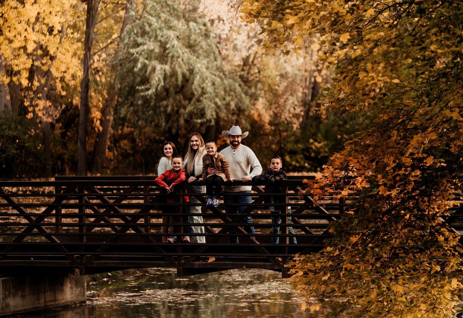 Family poses on wooden bridge surrounded by golden fall foliage in park setting.