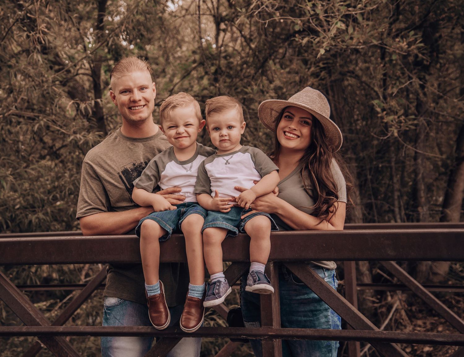 A family of four sits together on a rustic metal bridge in a wooded setting with warm sepia tones.
