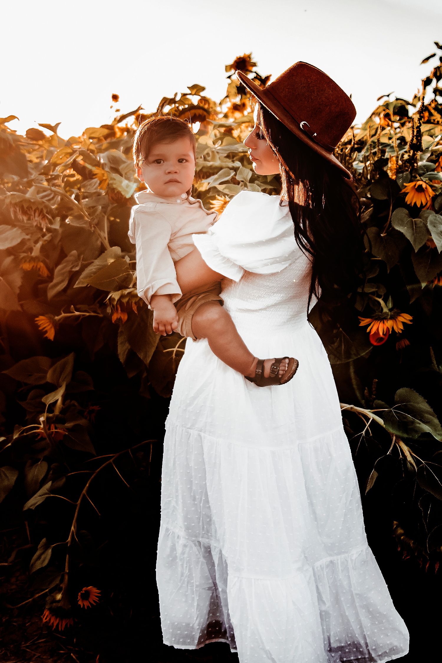 A sunset scene in a sunflower field with a white dress and brown hat silhouetted against the golden light.