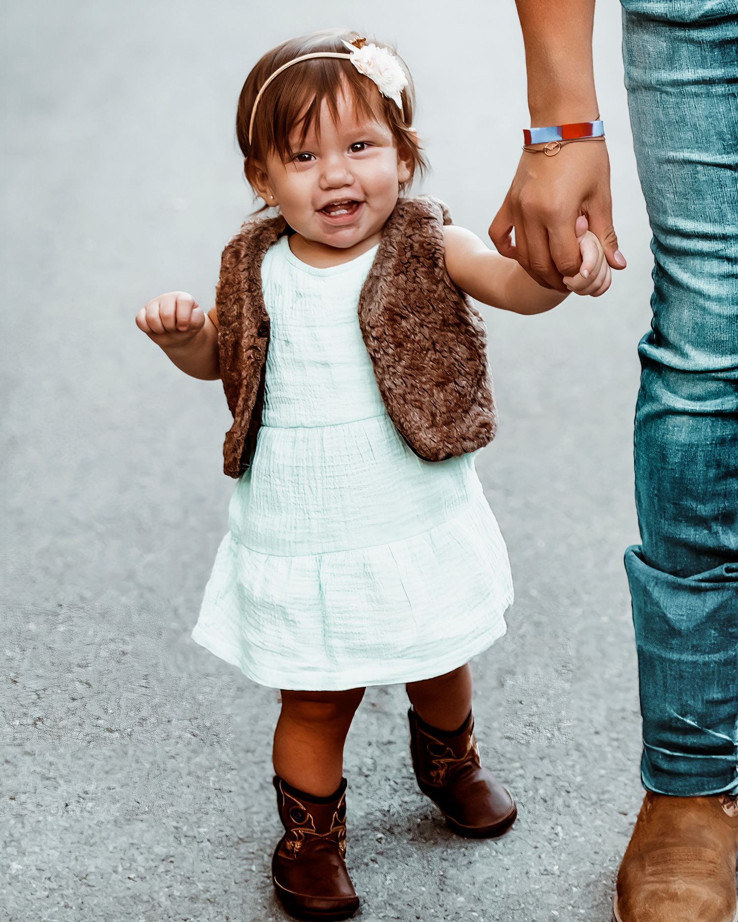 Small child in white dress and brown boots holds hands with adult wearing jeans and brown shoes.