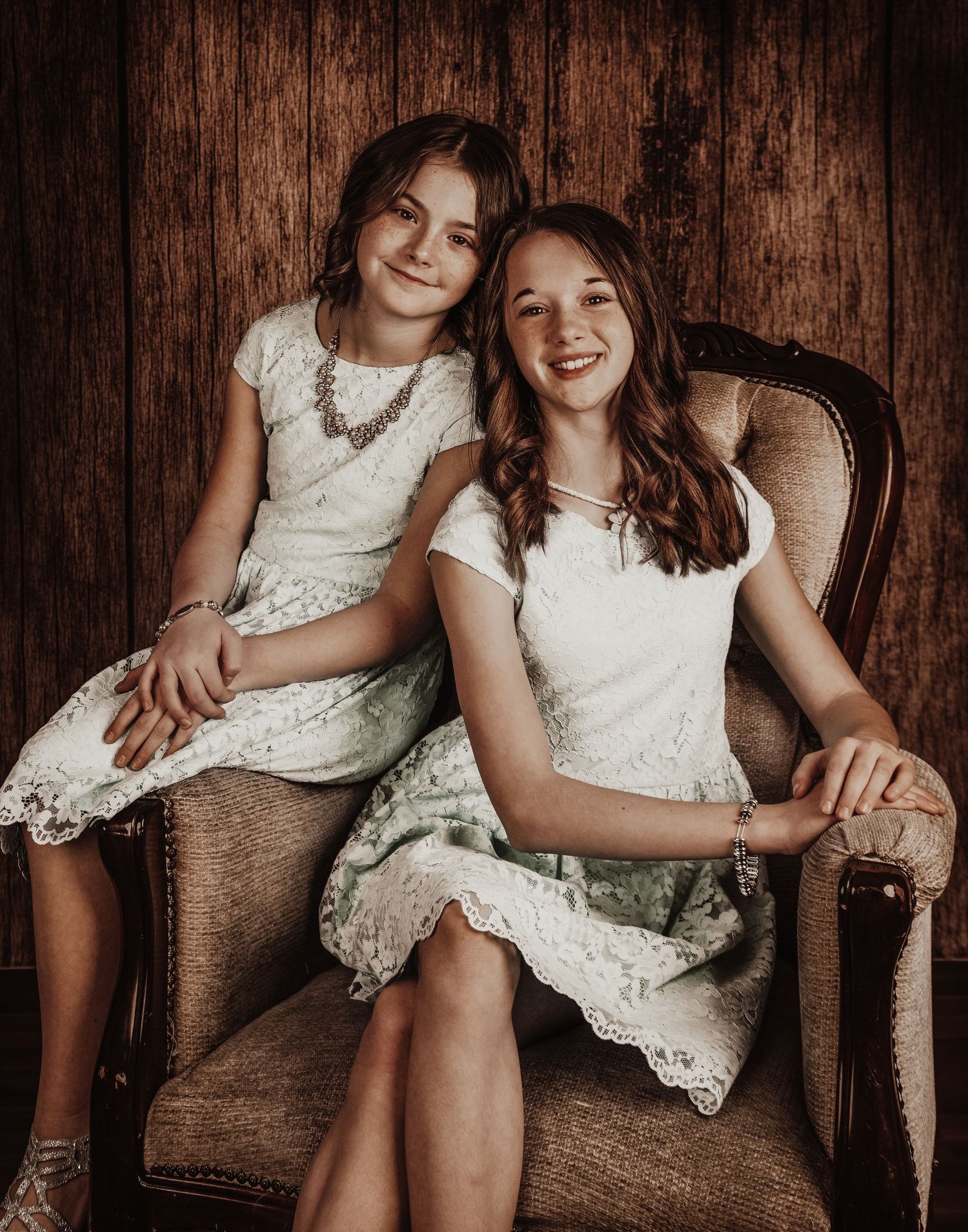 Two people in white lace dresses sitting together on a vintage chair against a rustic wooden wall background.