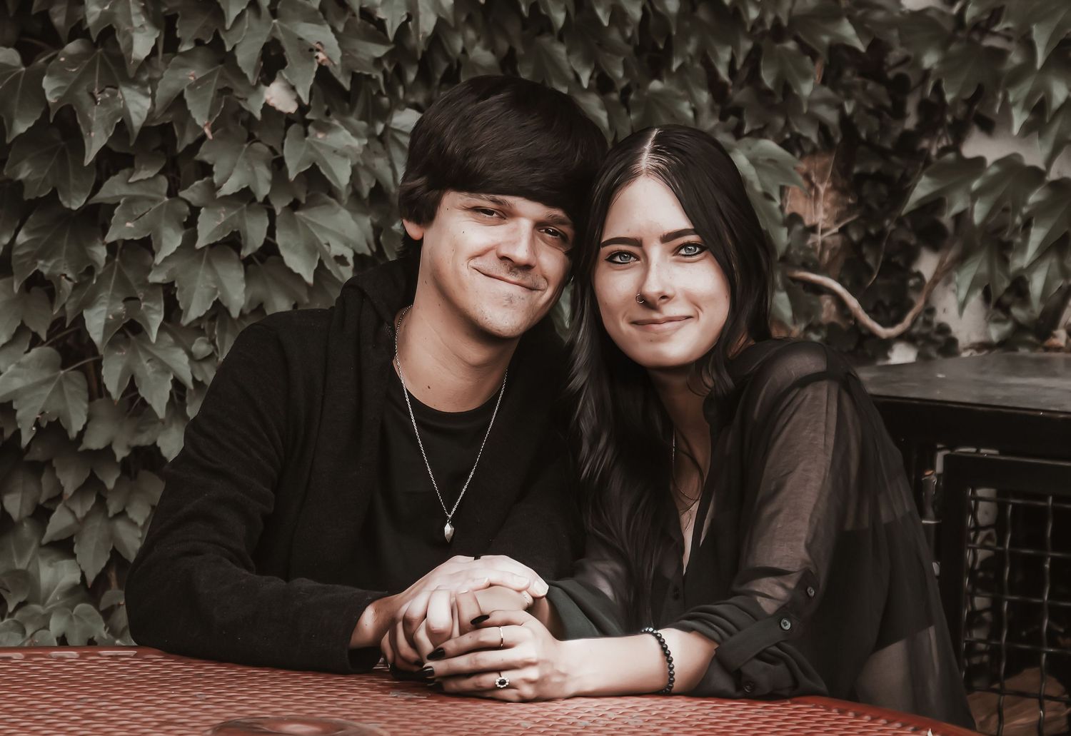 A couple sits close together at a red checkered table outdoors with lush green ivy in the background.