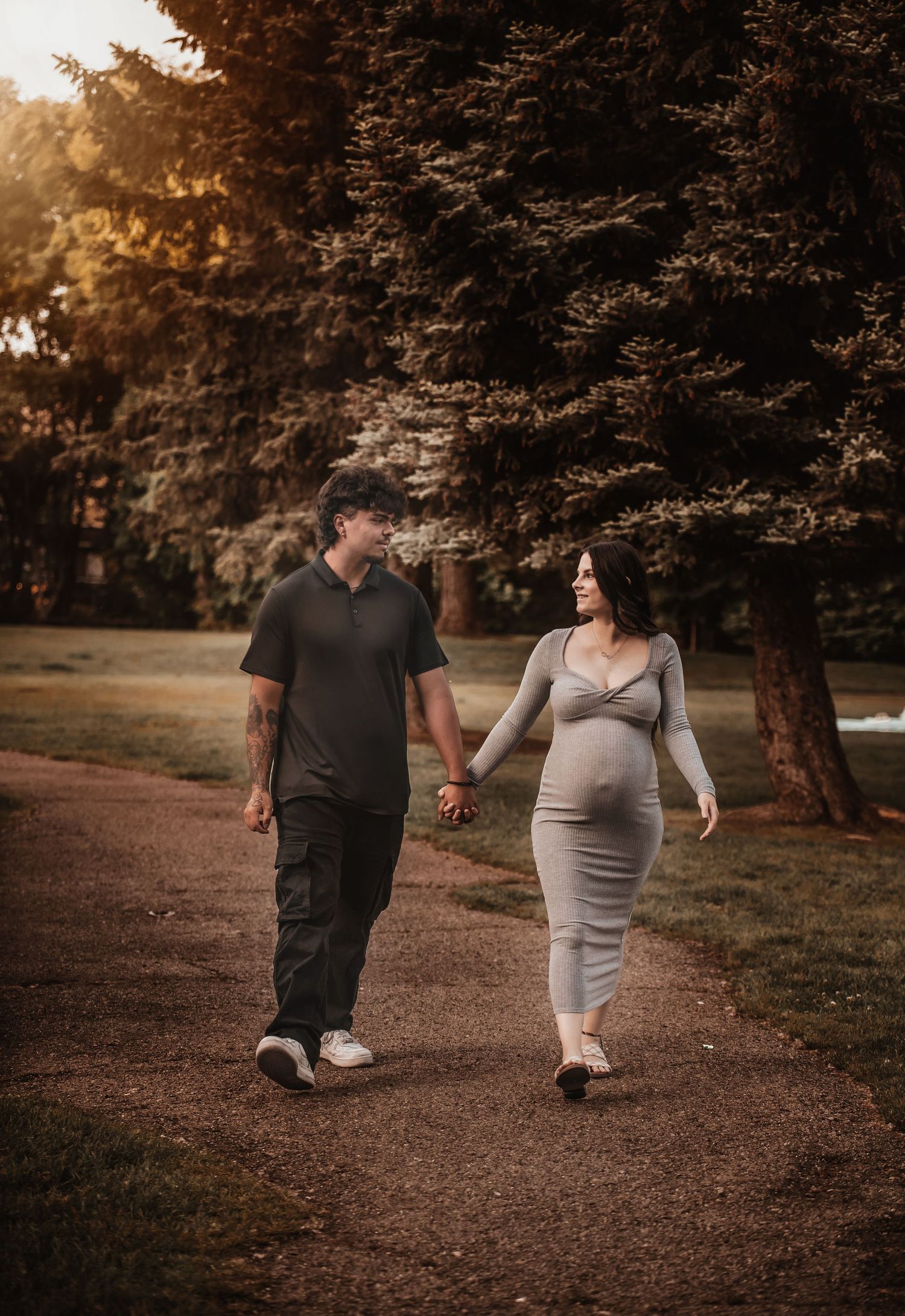 A couple in black and grey outfits walks together on a tree-lined path during golden hour sunset.