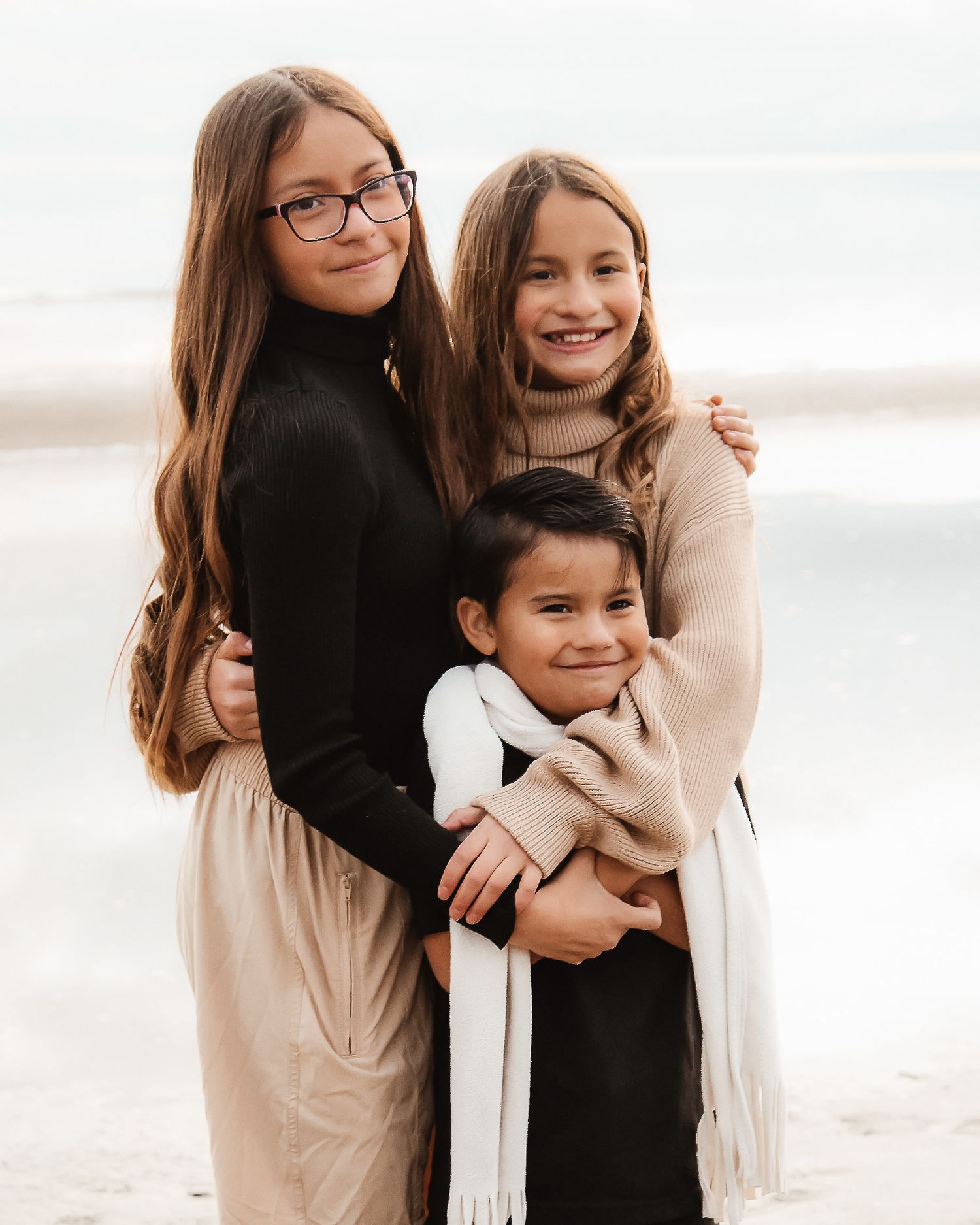 Three siblings in warm sweaters and scarves embrace while posing together on a misty beach.