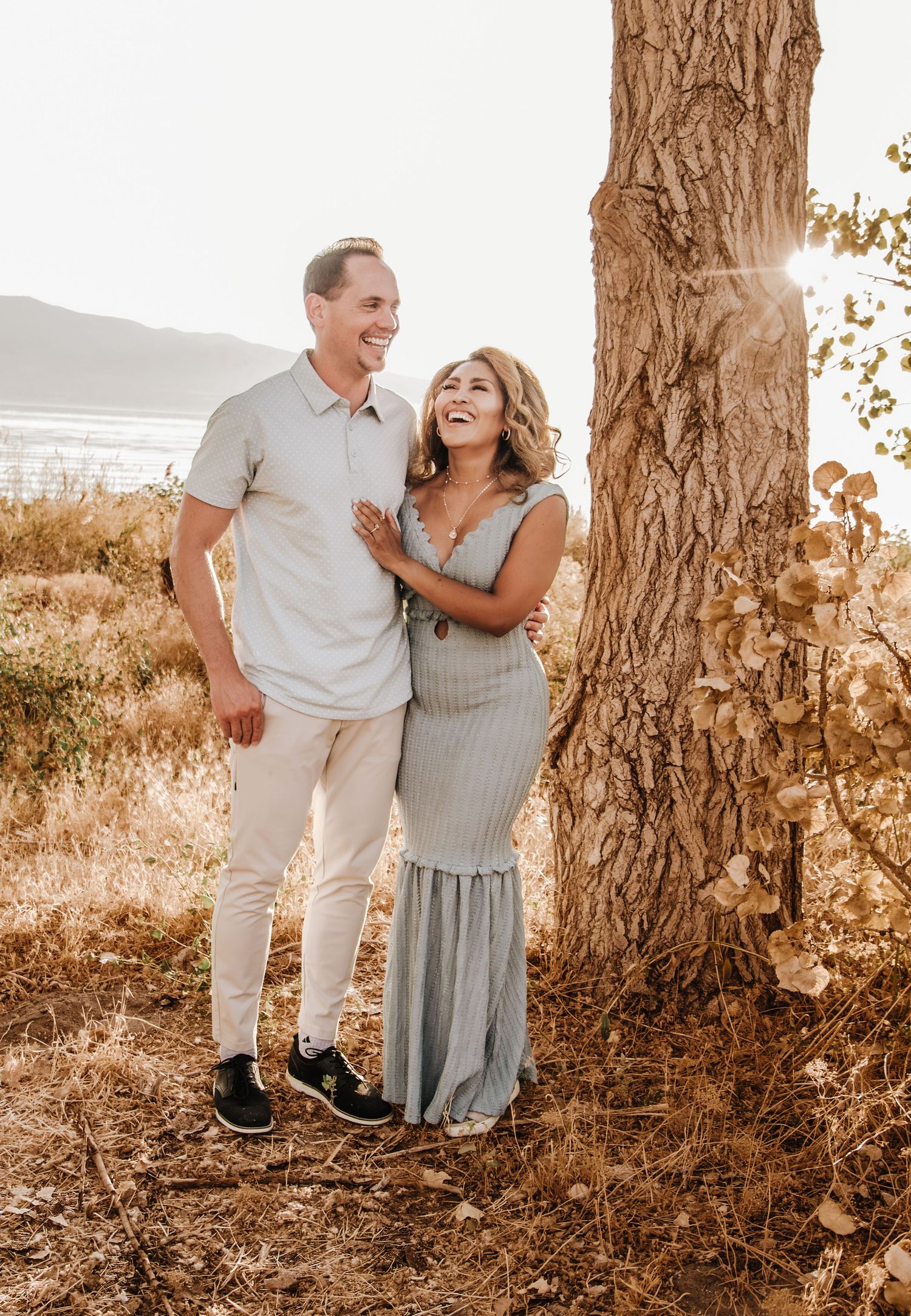 A couple in light colored outfits share intimate moments among tall trees during a warm sunset photoshoot.