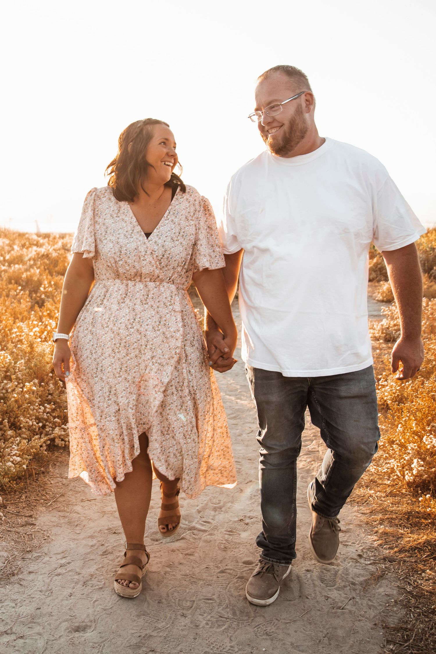 A couple walks hand in hand along a sunlit path, with the woman wearing a floral dress and man in casual attire.