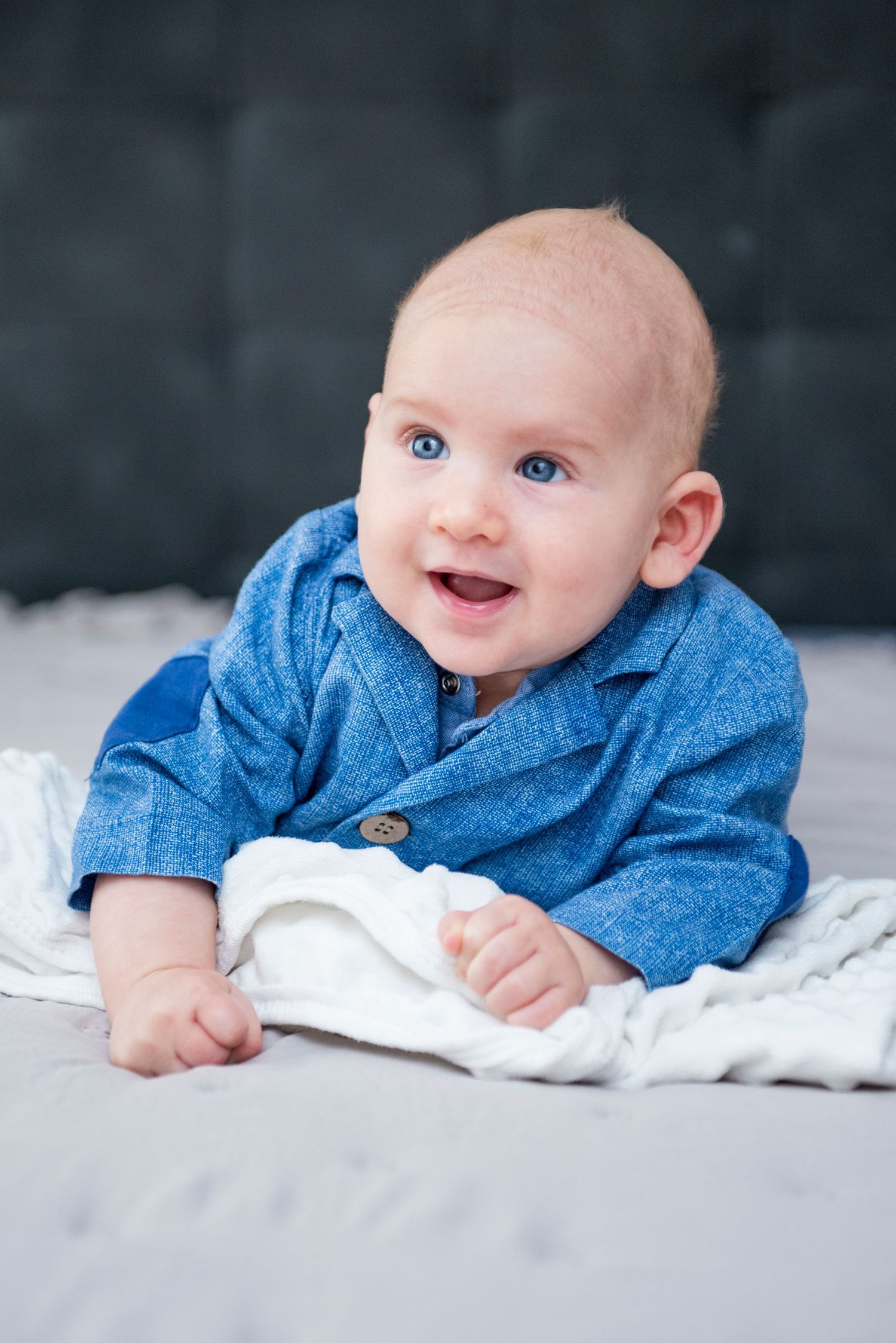 Baby in blue denim shirt lying on white blanket in studio setting with bright blue eyes and sweet smile.